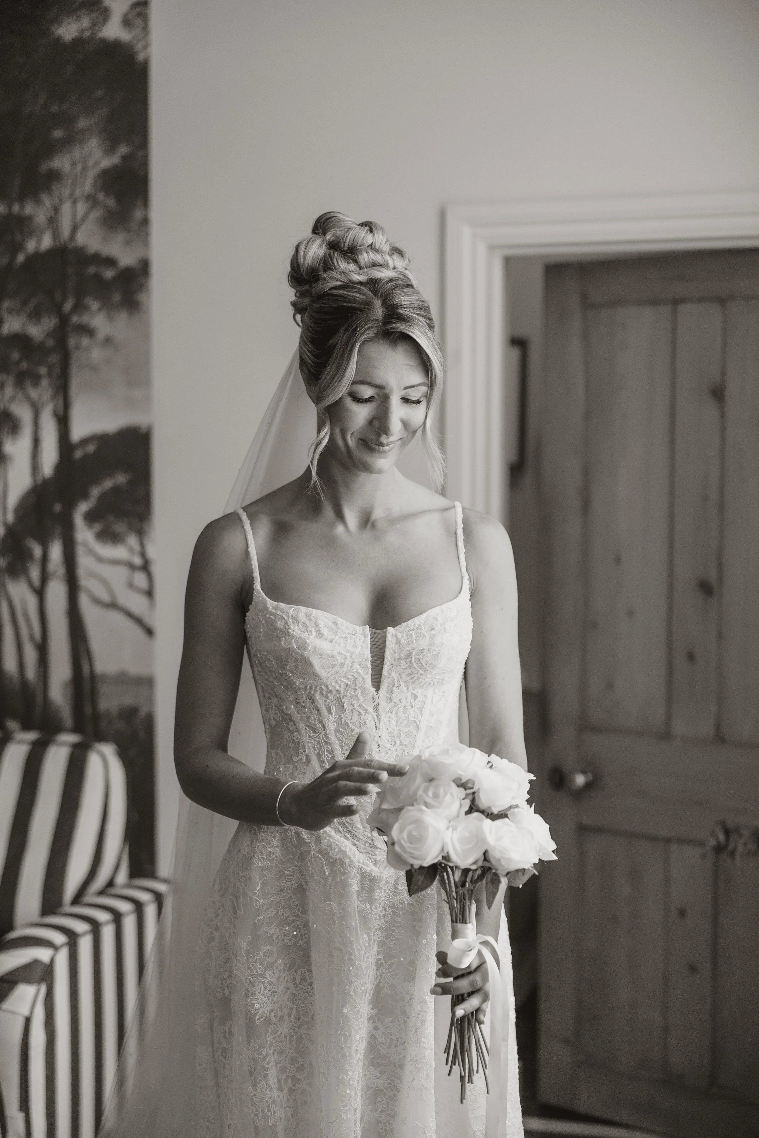 A bride in a lace wedding dress with spaghetti straps, holding a bouquet of white roses, standing indoors near a wooden door. She has her hair styled in an elegant updo with a veil, and is looking down with a gentle smile.