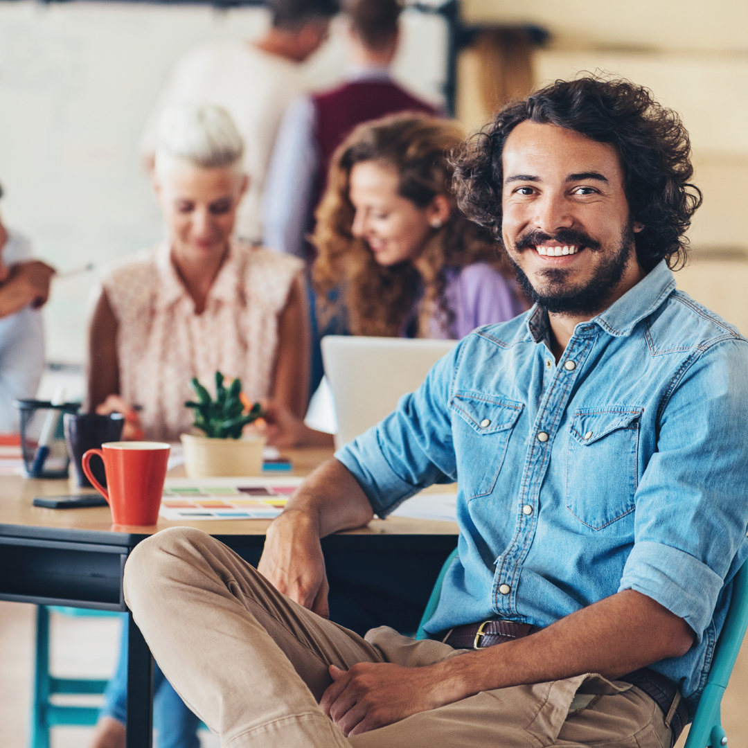 A group of people in a meeting room, with a smiling man in a denim shirt sitting at the front and others working and chatting in the background.