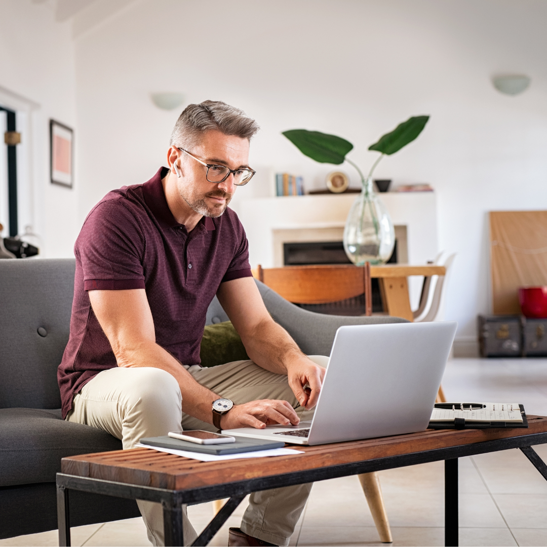 A man with glasses and a beard working on a laptop in a modern living room