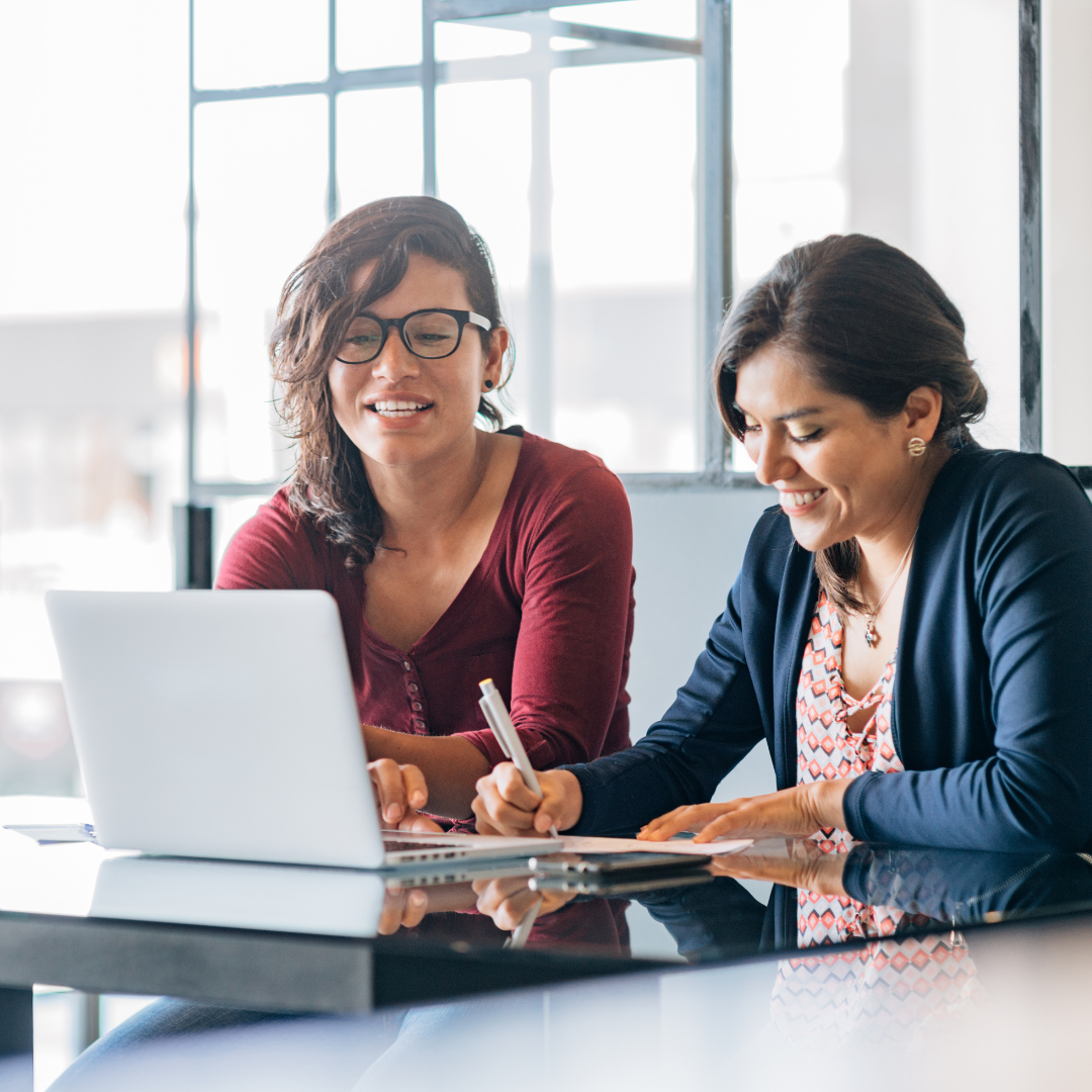 Two women sitting at a table working on a laptop together, one is writing on a notepad.