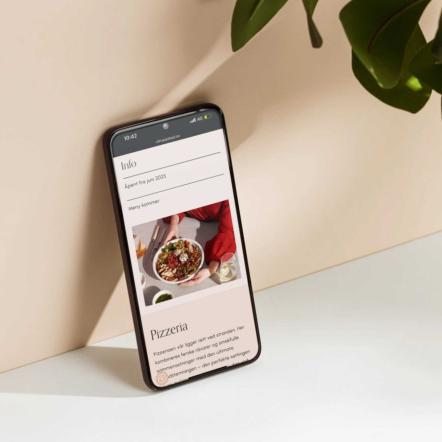 En mobil som viser nettsiden til Vilma Sjøbad i responsivt format. Mobilen lener seg på en dus rosa vegg ved siden av en grønn plante.