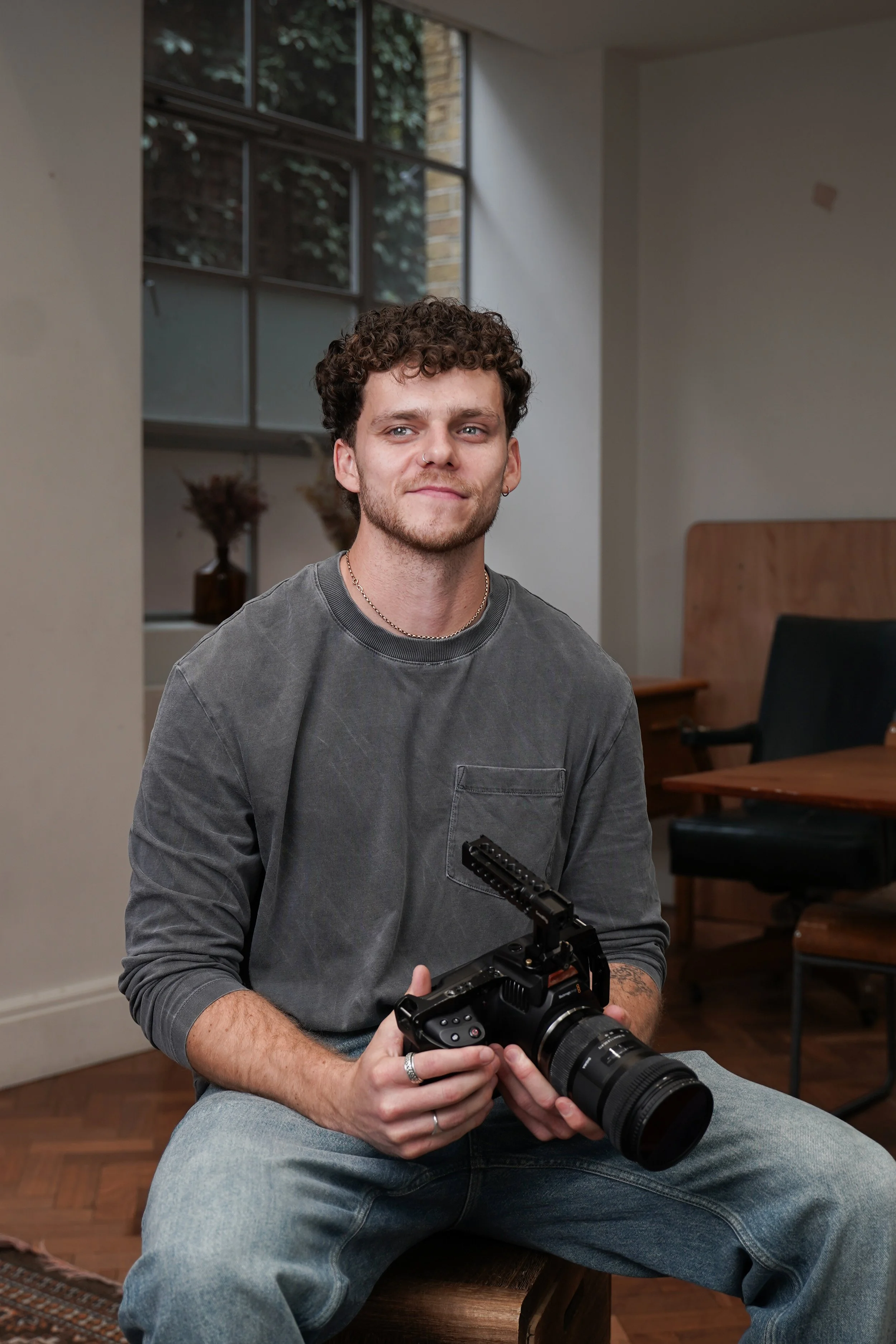A young man with curly hair, facial hair, and wearing a nose ring, sitting indoors and holding a professional camera with a large lens. He is wearing a gray long-sleeve shirt and blue jeans. The room has large windows, a wooden table, and chairs in the background.