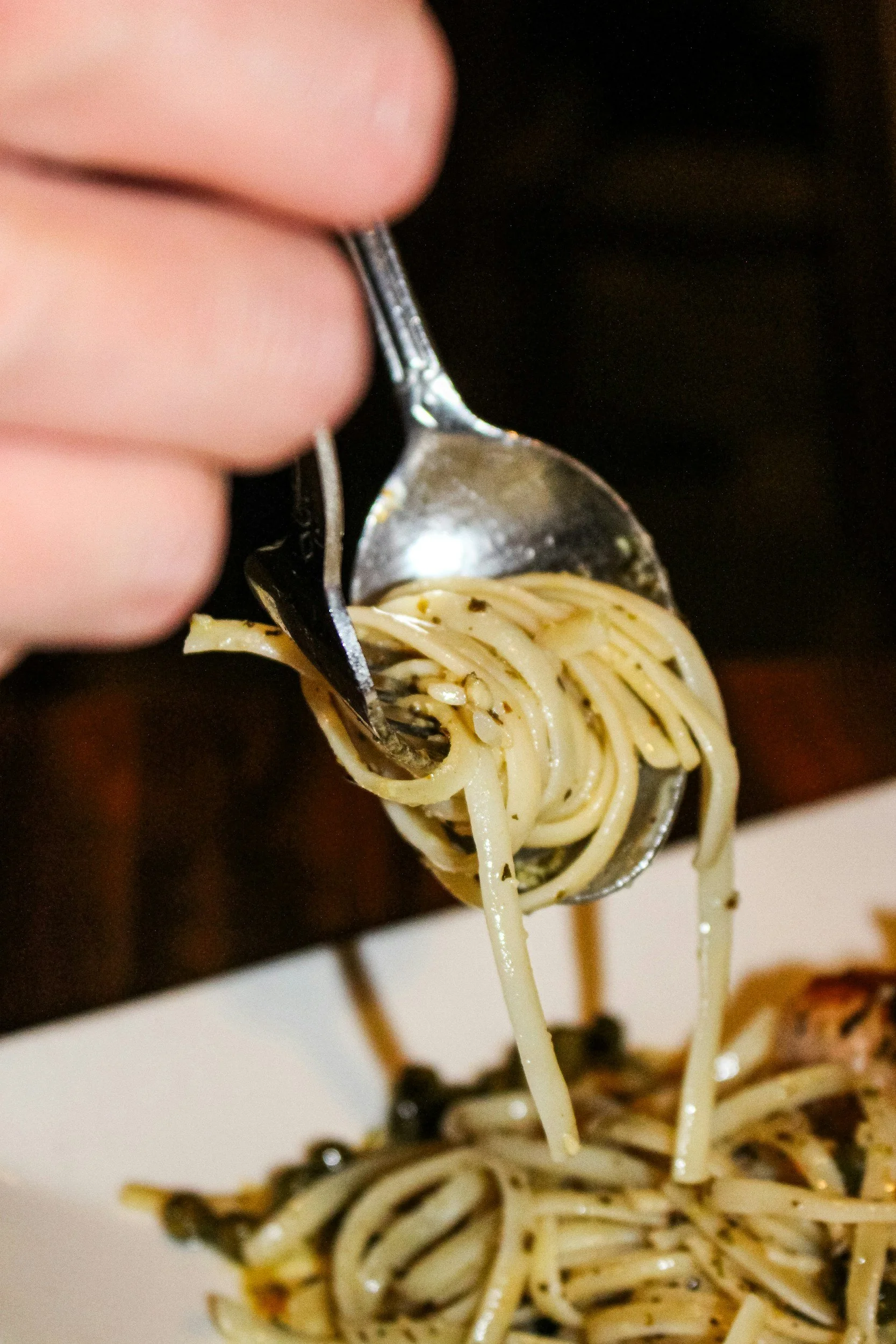 Close-up of a fork lifting spaghetti noodles with black pepper and herbs, with a dark background.