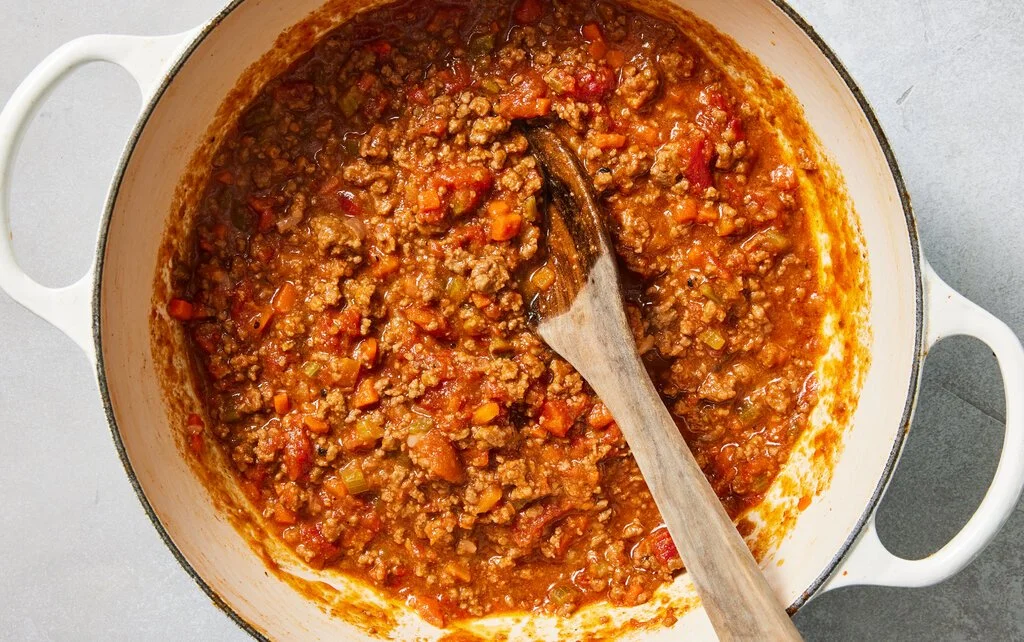 A pot of cooked ground beef chili with vegetables like tomatoes and celery, with a wooden spoon inside.