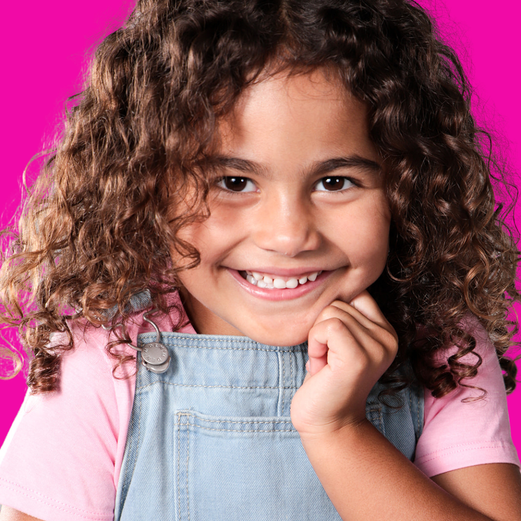 Close-up of a smiling young girl with curly brown hair, wearing a pink shirt and denim overalls, against a pink background.