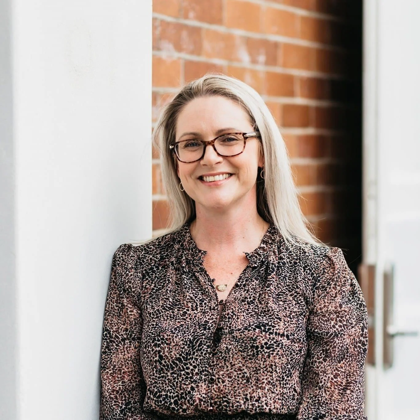 A smiling woman with long blonde hair, wearing glasses and a leopard print blouse, leaning against a white wall with a brick wall in the background.