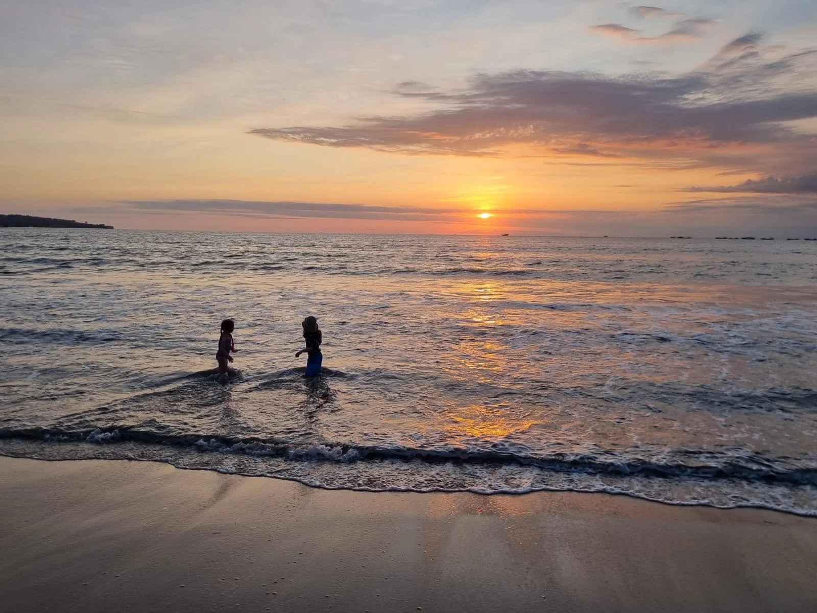 Two children playing in the ocean at sunset with a colorful sky and calm waves.