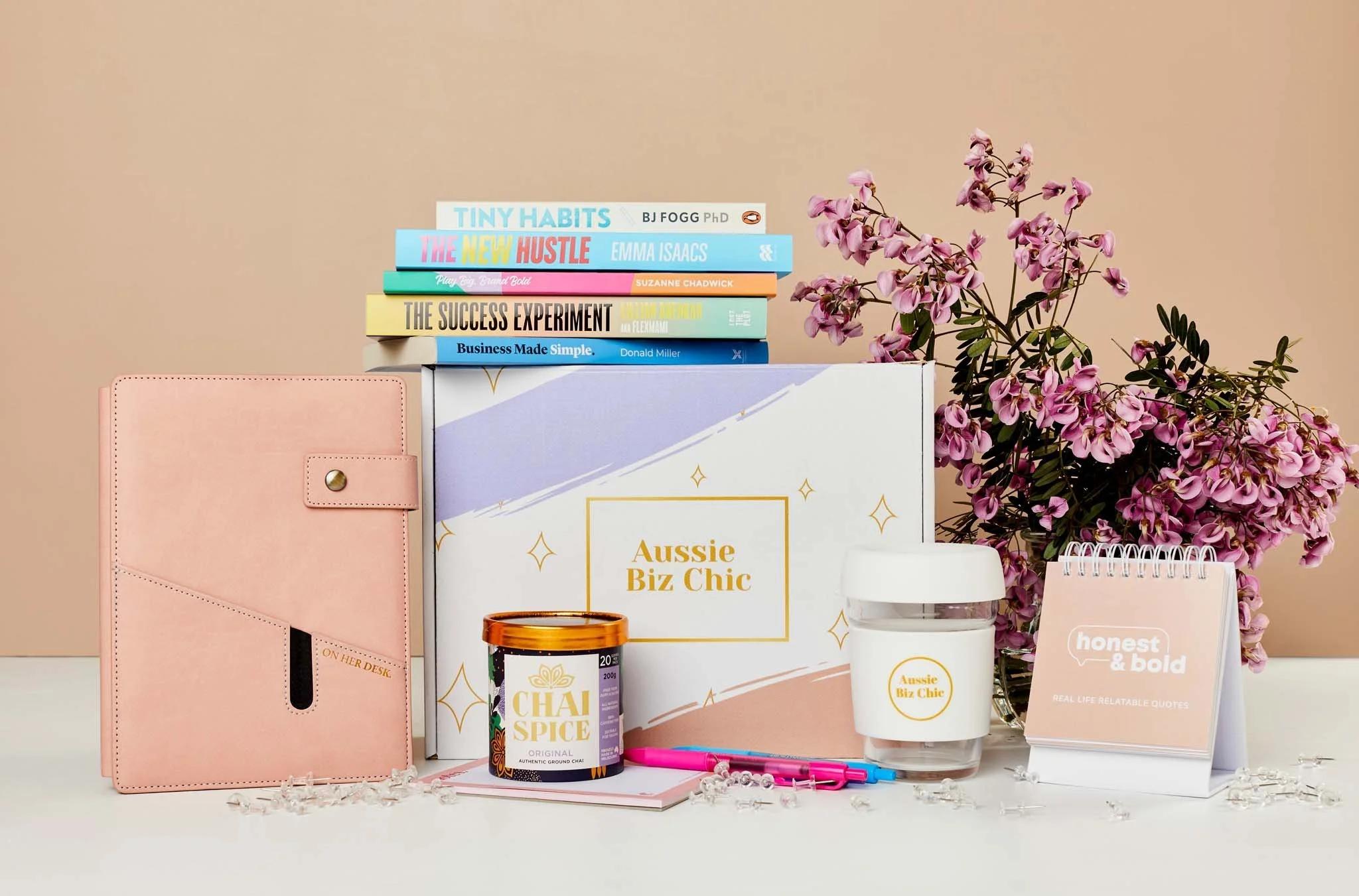 Pink notebook, stack of books, pink flowers, tea container, glass with 'Aussie Biz Chic' label, pink and blue pens, and a pink notepad with 'honest & bold' on it, arranged on a white surface with a beige background.
