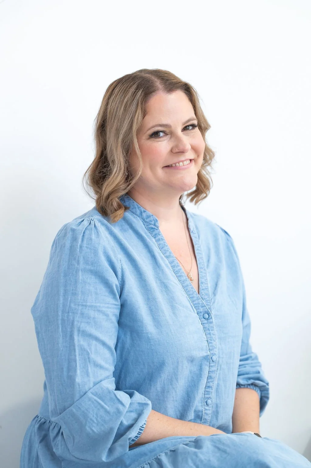 A woman with shoulder-length brown hair, wearing a light blue button-up shirt, smiling against a white background.