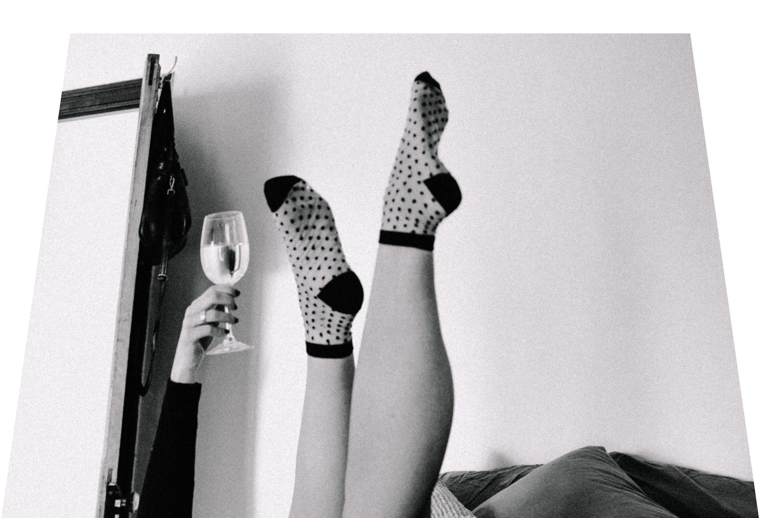 A black and white image of a womens feet up in the air. She is wearing socks and holding a glass of wine up in the air too.