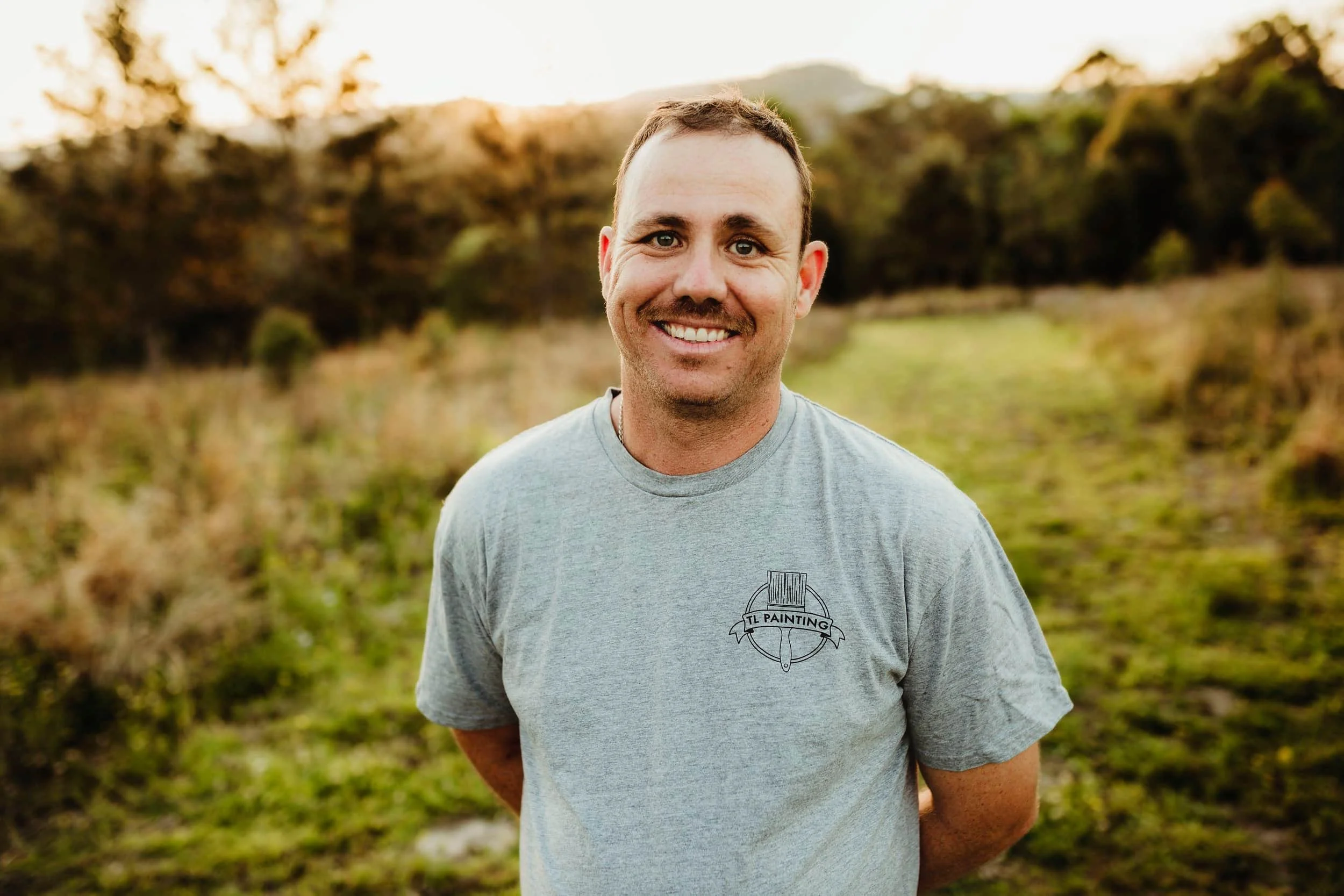 A smiling man standing outdoors in a grassy field during sunset, wearing a gray T-shirt with a logo that reads 'TL Painting'.