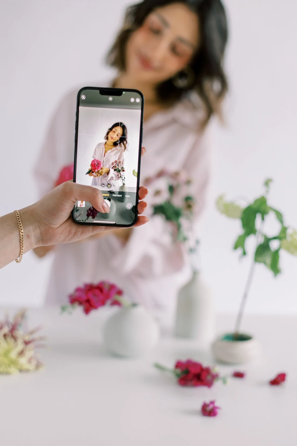 A woman with short dark hair and hoop earrings poses with flowers in a white vase as another person takes her photo with a smartphone, with the focus on the phone screen.