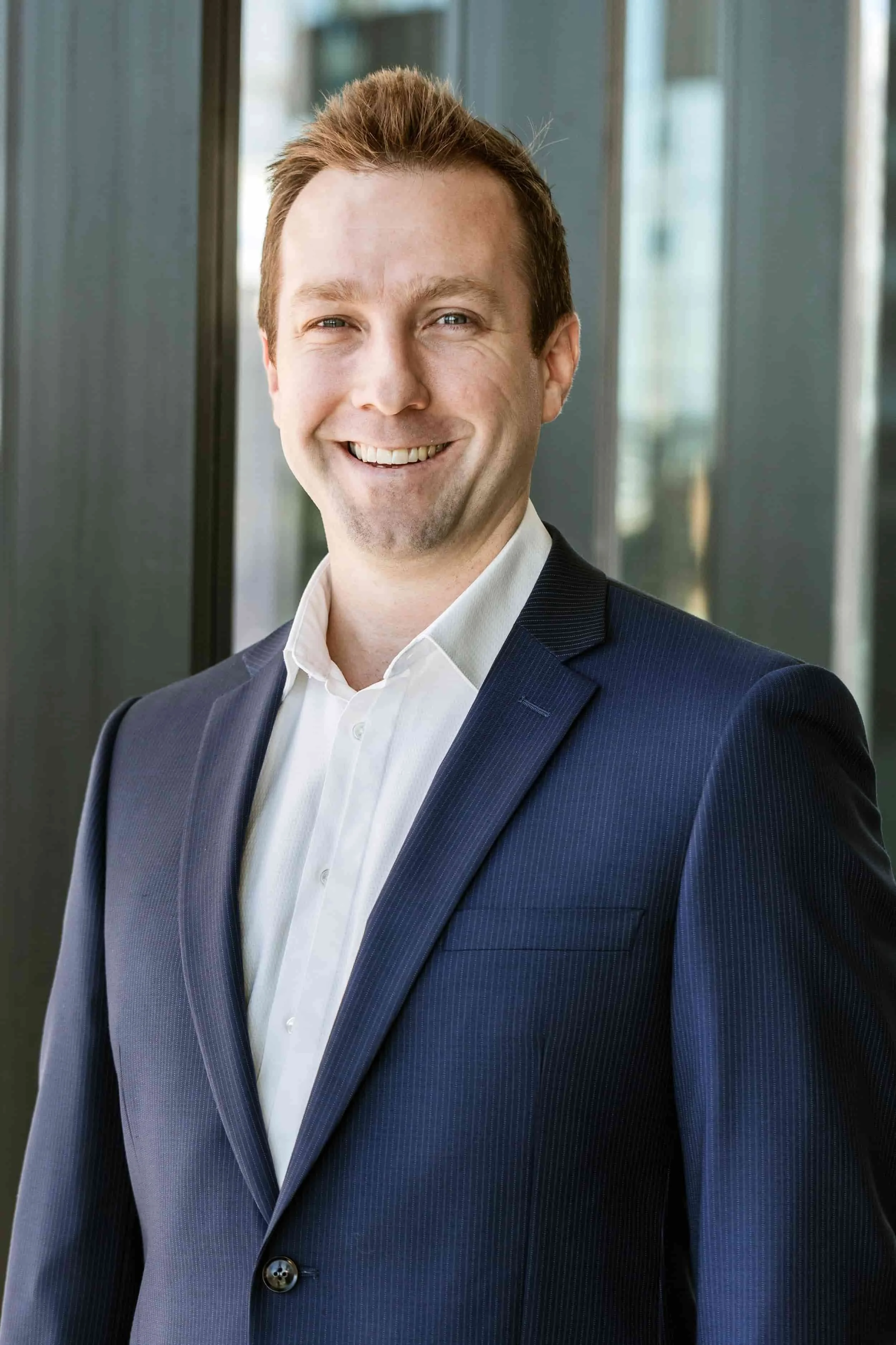 A smiling man in a dark blue suit and white shirt standing indoors near large windows.