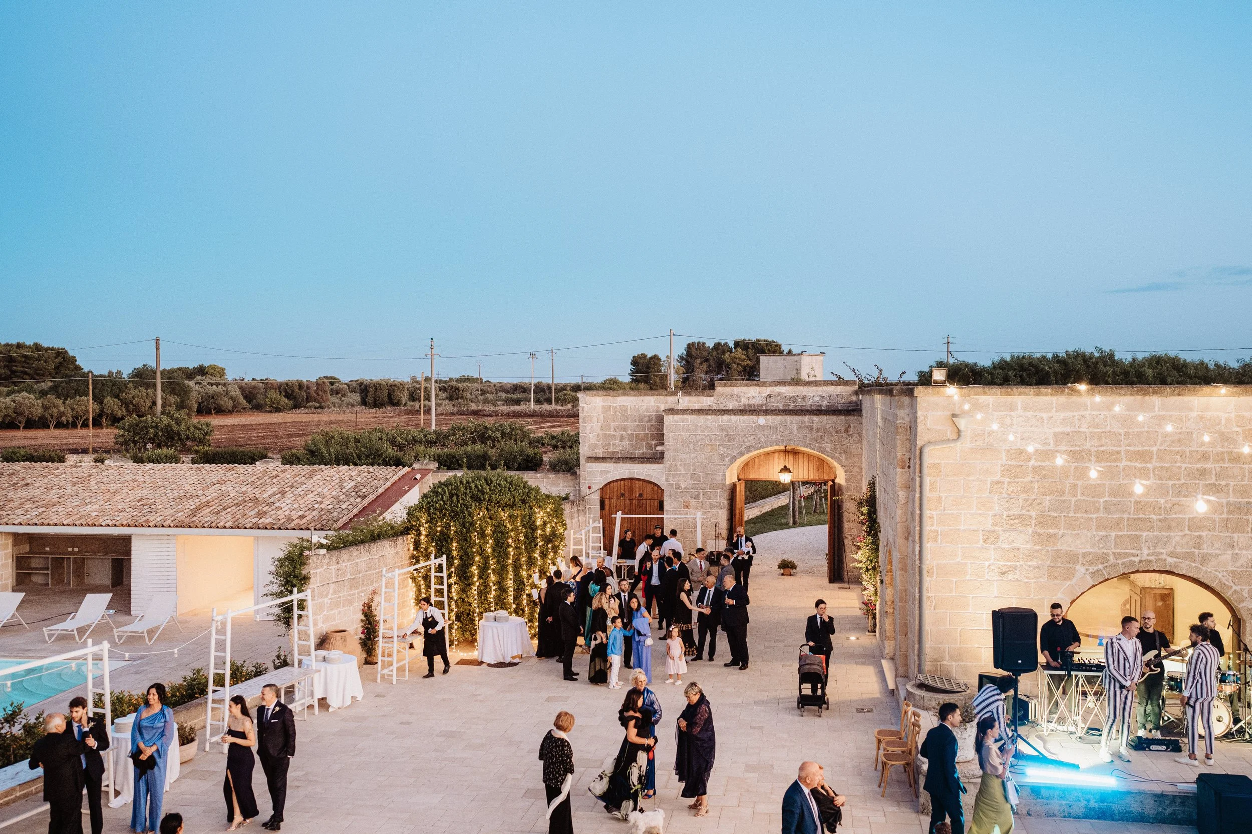 Outdoor evening event at a rustic venue with people socializing, a live band playing, and string lights decorating the area.