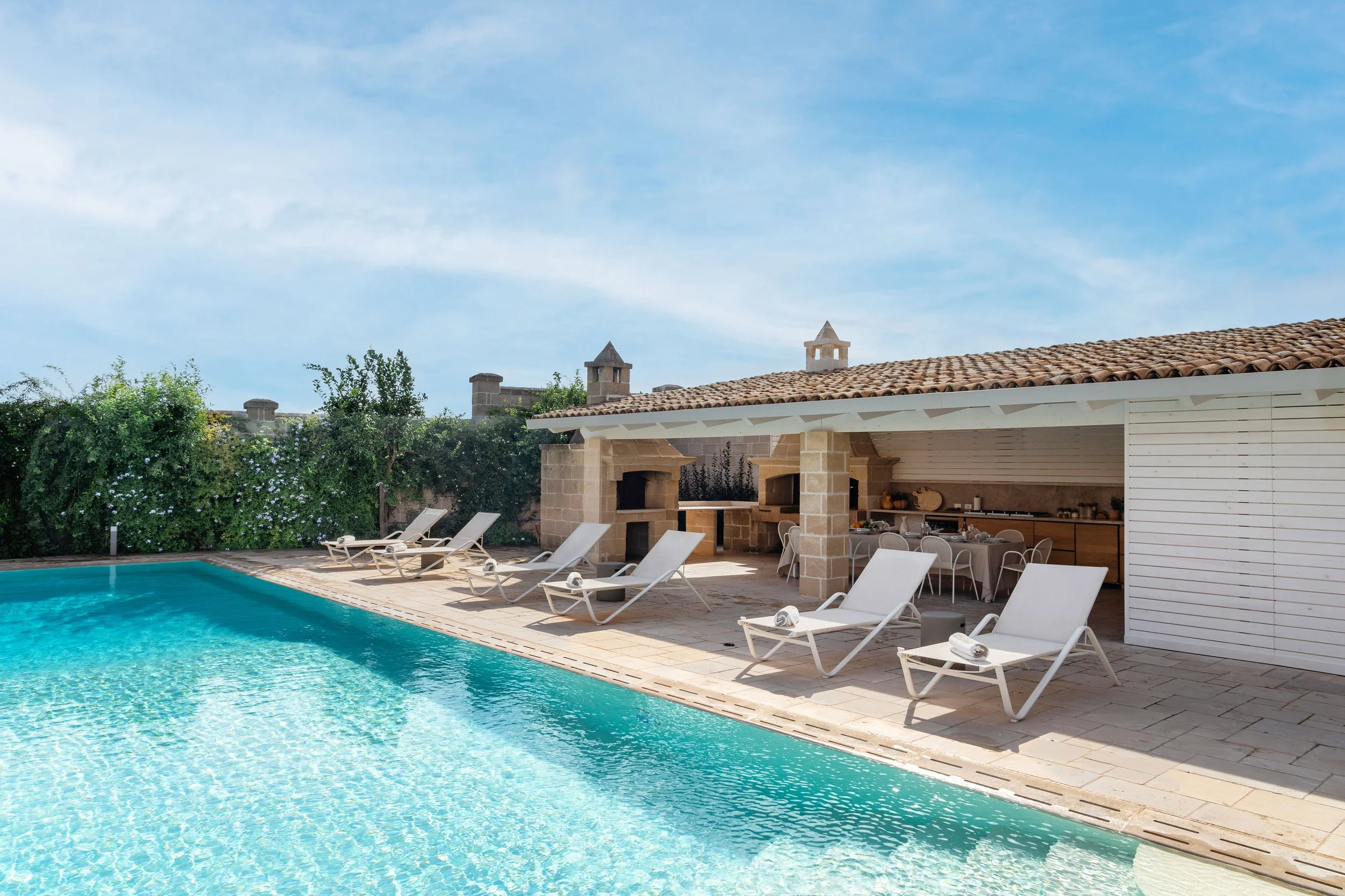 Outdoor swimming pool with sun loungers and a covered patio area, surrounded by greenery.
