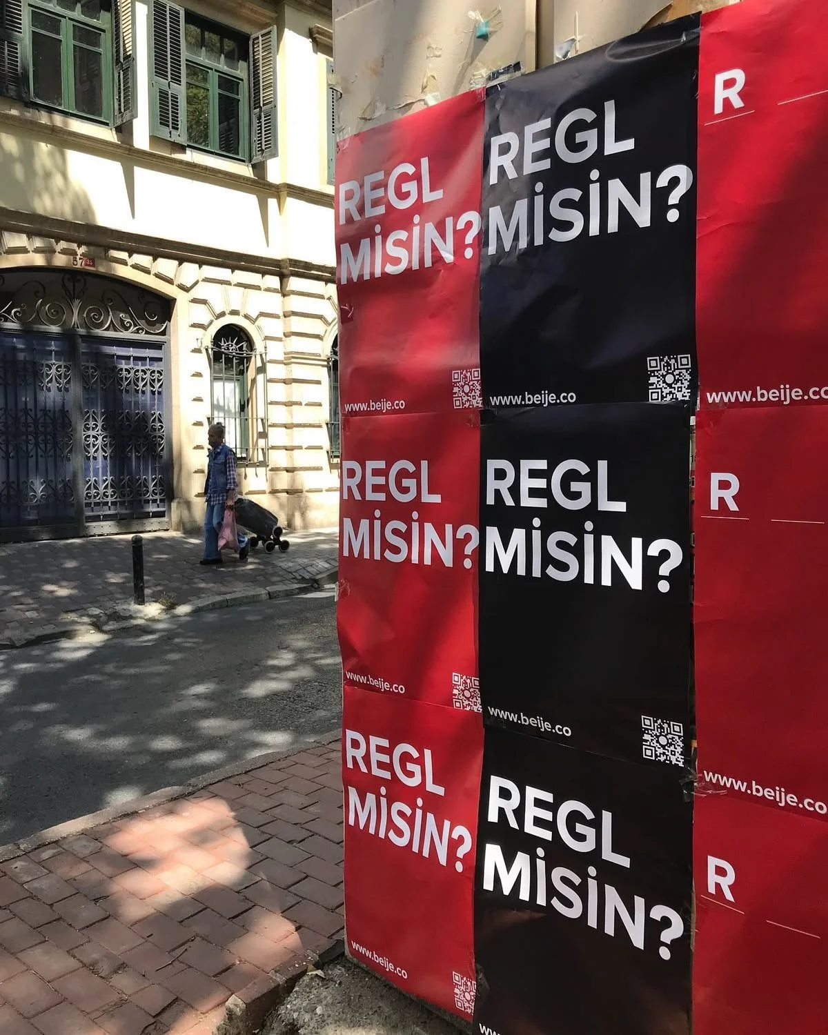 A large red and black poster on a city sidewalk displays the Turkish words 'REGL MISIN?' repeated multiple times. In the background, a man wearing blue clothing and a head covering walks along the street, pulling a shopping cart.
