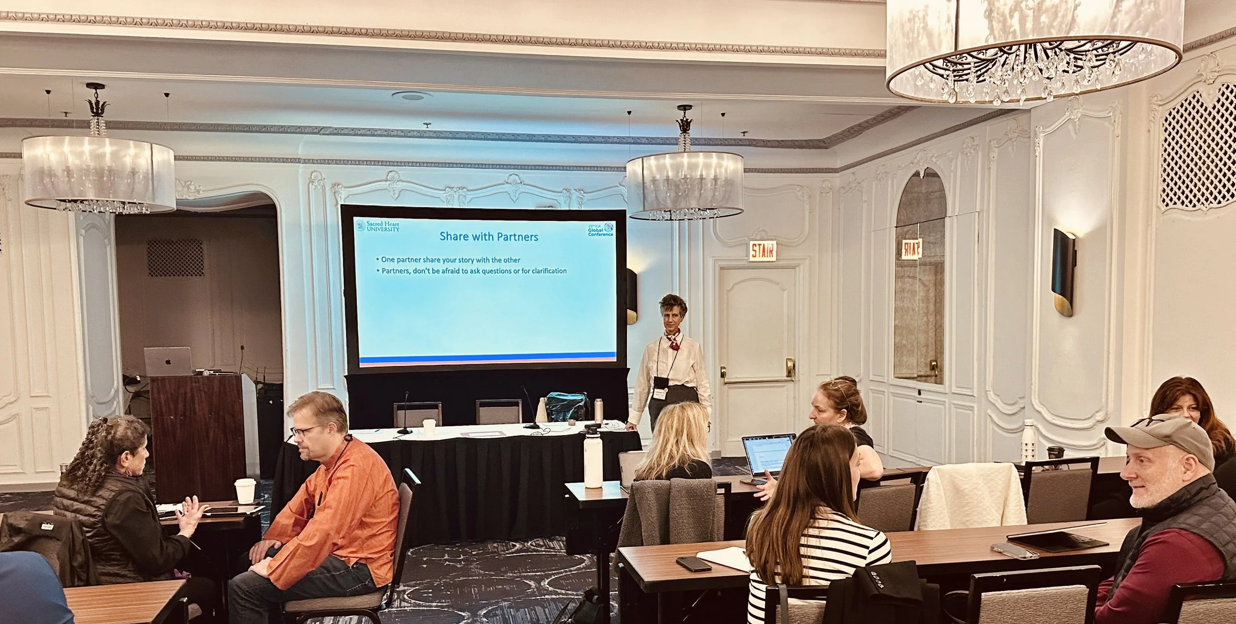 groups of two people sitting in pairs at spaced out tables with one person at the front of the room with screen behind them with "Share with Partner" prompt on it.