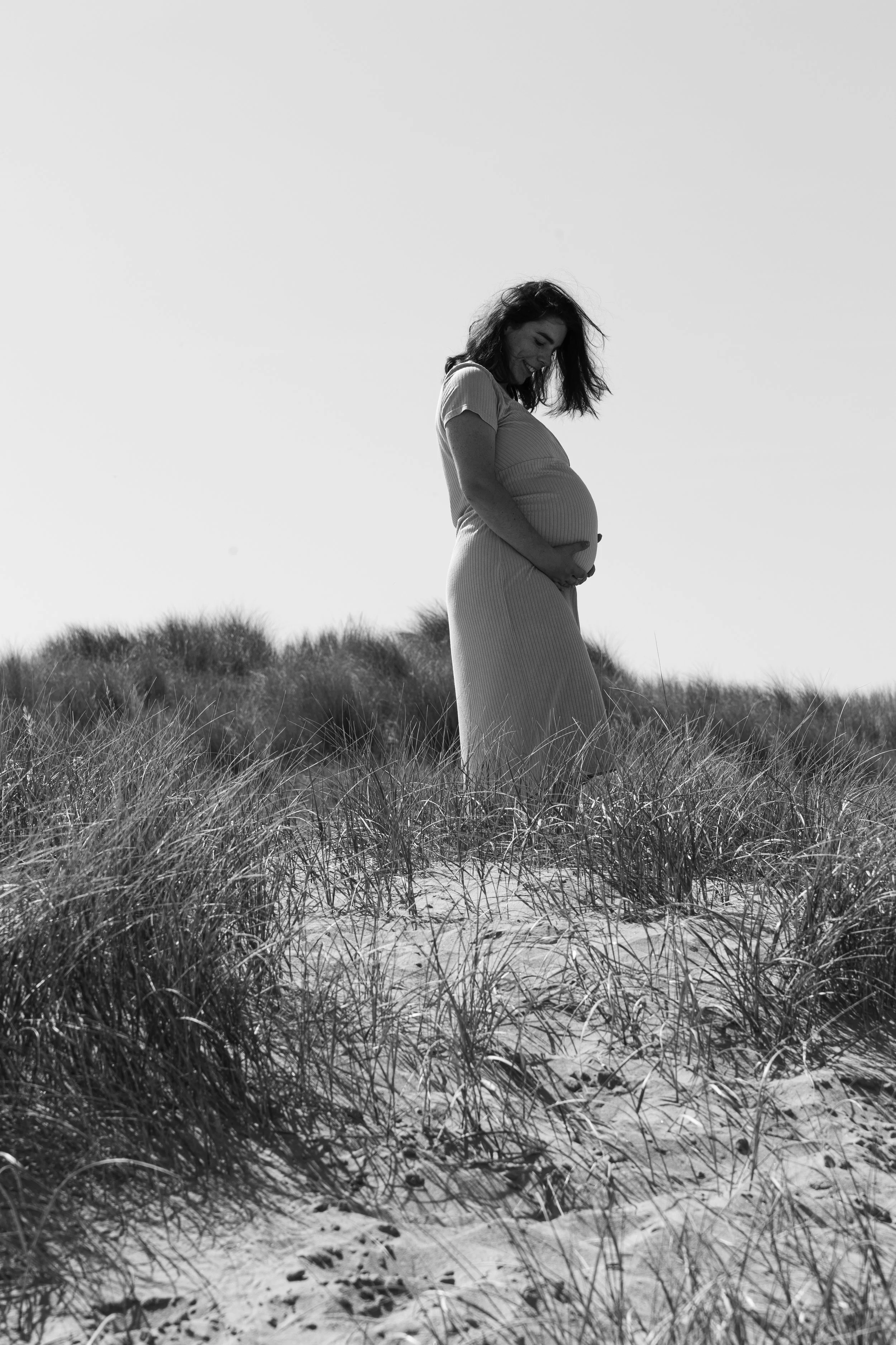Een zwangere vrouw staat in een duinachtig gebied met gras, lachend en met haar handen op haar buik.