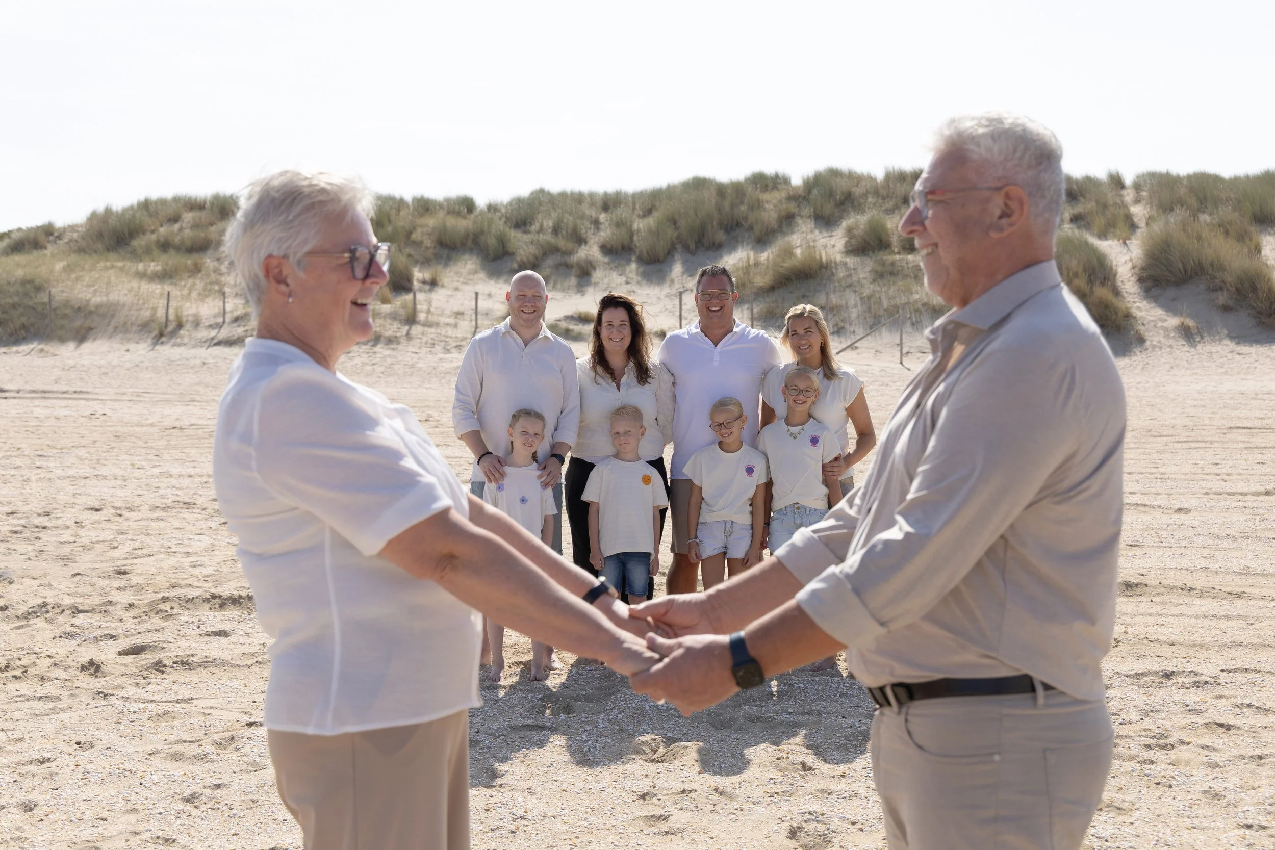 Twee oudere mensen handen schudden op het strand met een groep volwassenen en kinderen op de achtergrond.