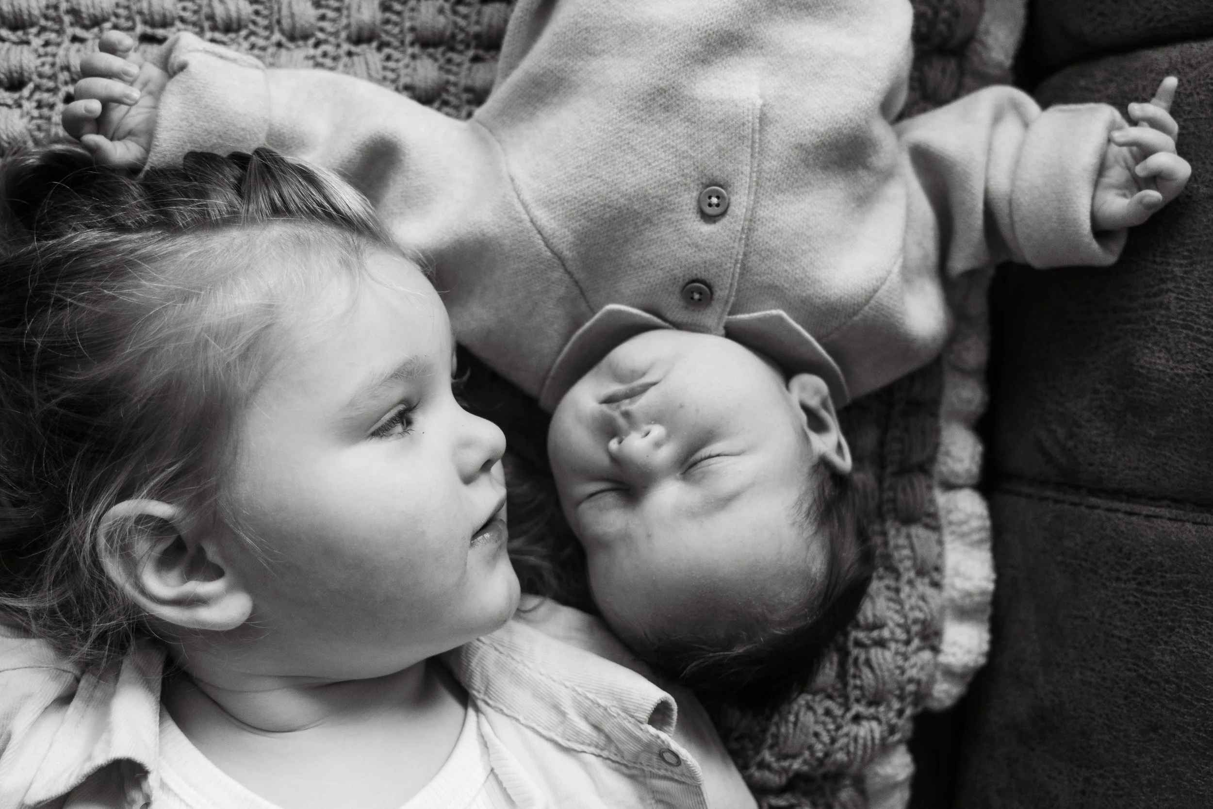 Twee kinderen liggen naast elkaar op de grond, één met blik naar rechts en het andere met ogen dicht, beide op een tapijt.
