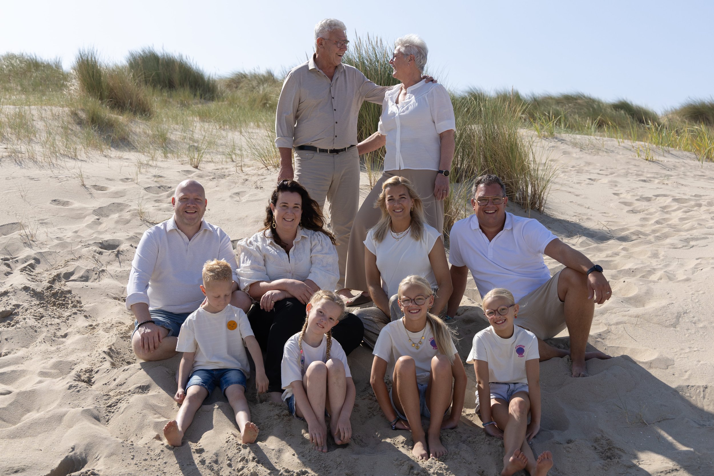 Een groep mensen, bestaande uit volwassenen en kinderen, zit en staat op het zand van een duinstrand. Ze lachen en genieten van de zonnige dag, met gras en duinen op de achtergrond.