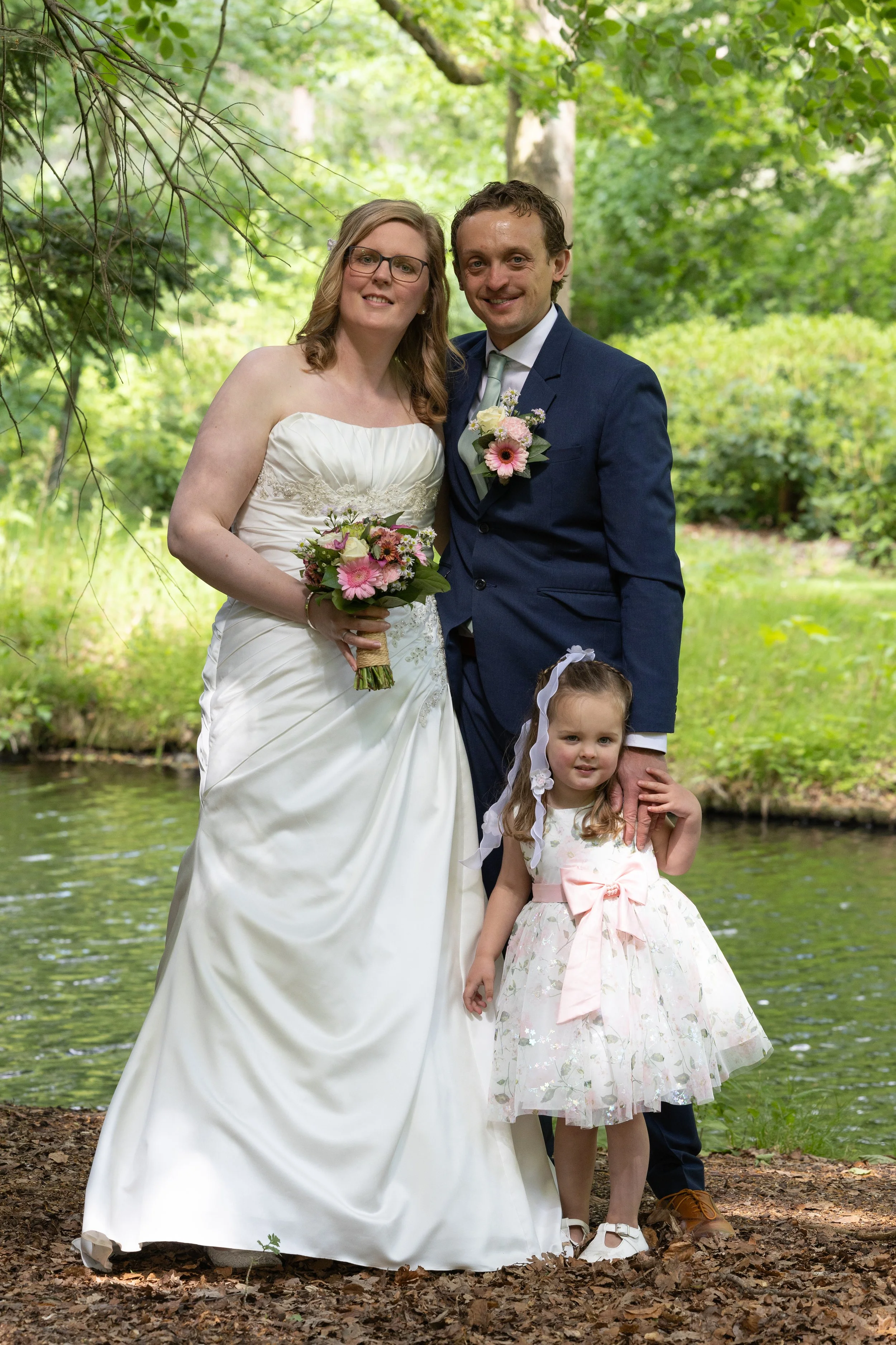 Een stel in huwelijkskleding staat aan de rand van een meer in een bosrijke omgeving, met een klein meisje in een lichtroze jurk met een grote strik voor zich, de vrouw houdt een bloemenboeket vast.