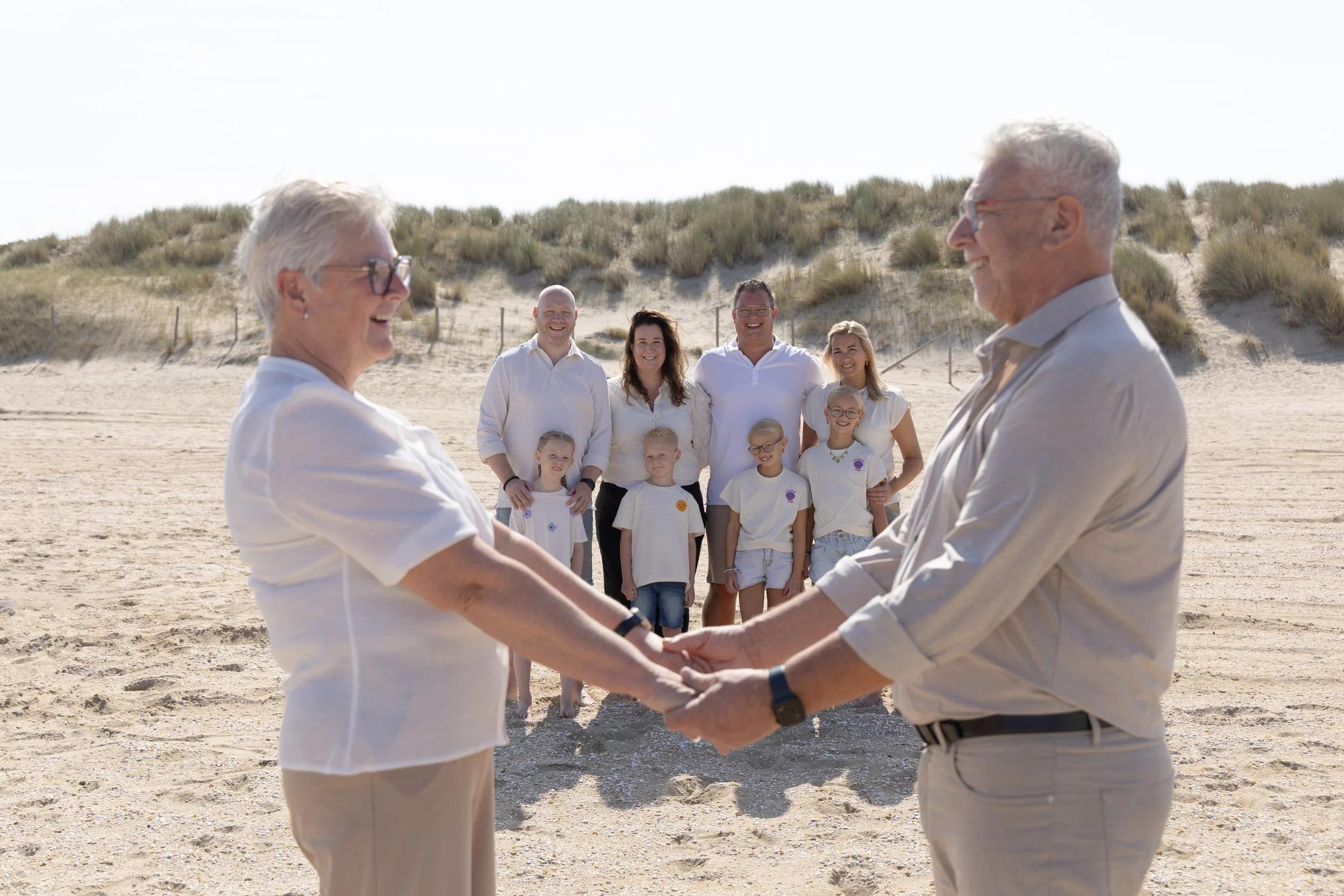 Een groep van tien mensen, waaronder ouderen en kinderen, op een strand, die betrokken zijn bij een afscheidsceremonie waarbij twee mannen elkaar de hand schudden en de rest toekijkt, allemaal gekleed in lichte kleuren.