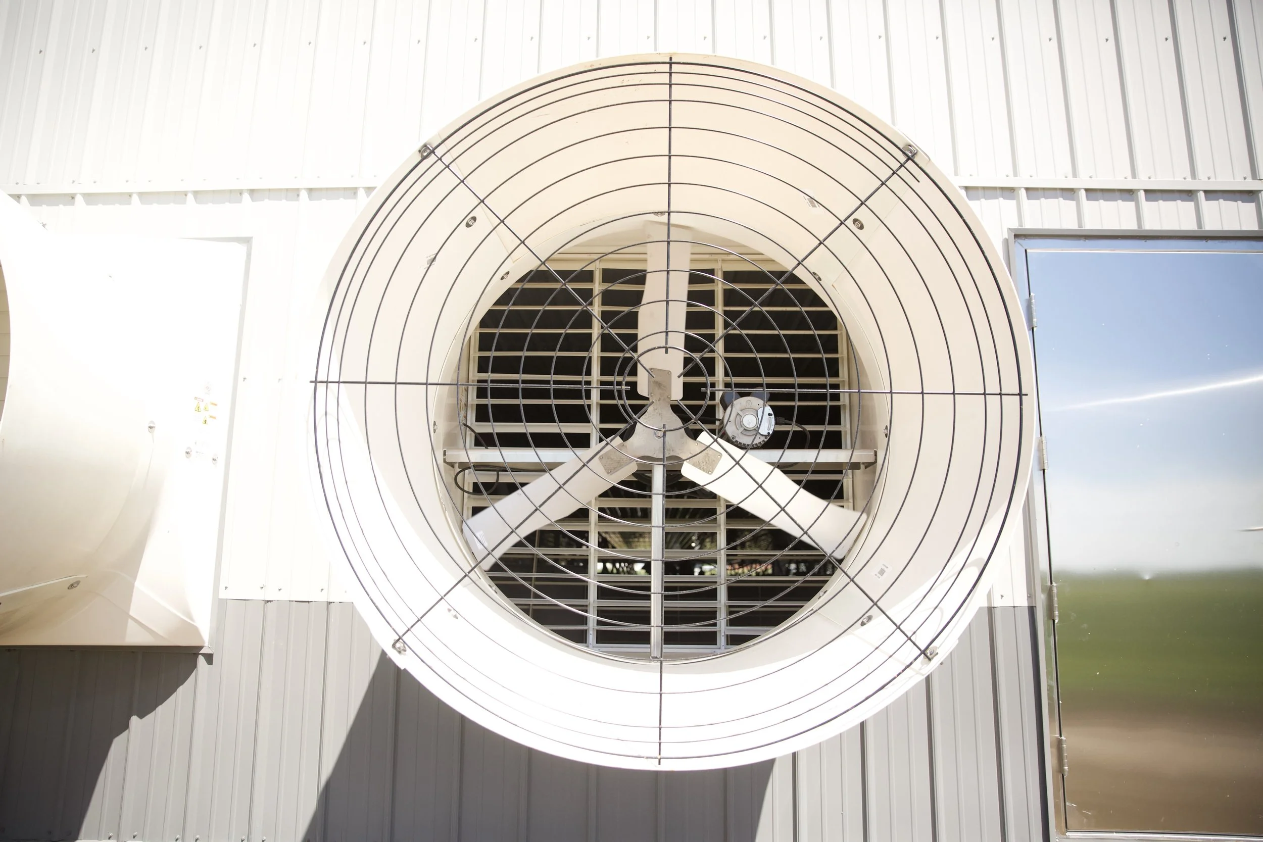 Large industrial vent or exhaust fan mounted on a white building exterior wall with metal siding, against a clear blue sky.