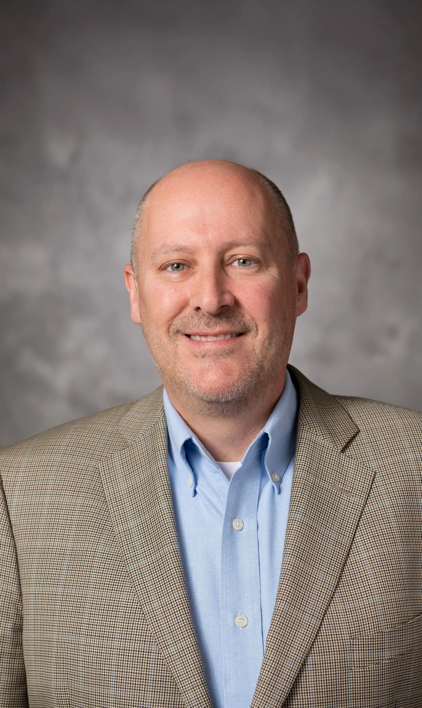 Portrait of a smiling middle-aged man with light skin, a shaved head, and a goatee, wearing a beige checkered blazer and a light blue button-up shirt.