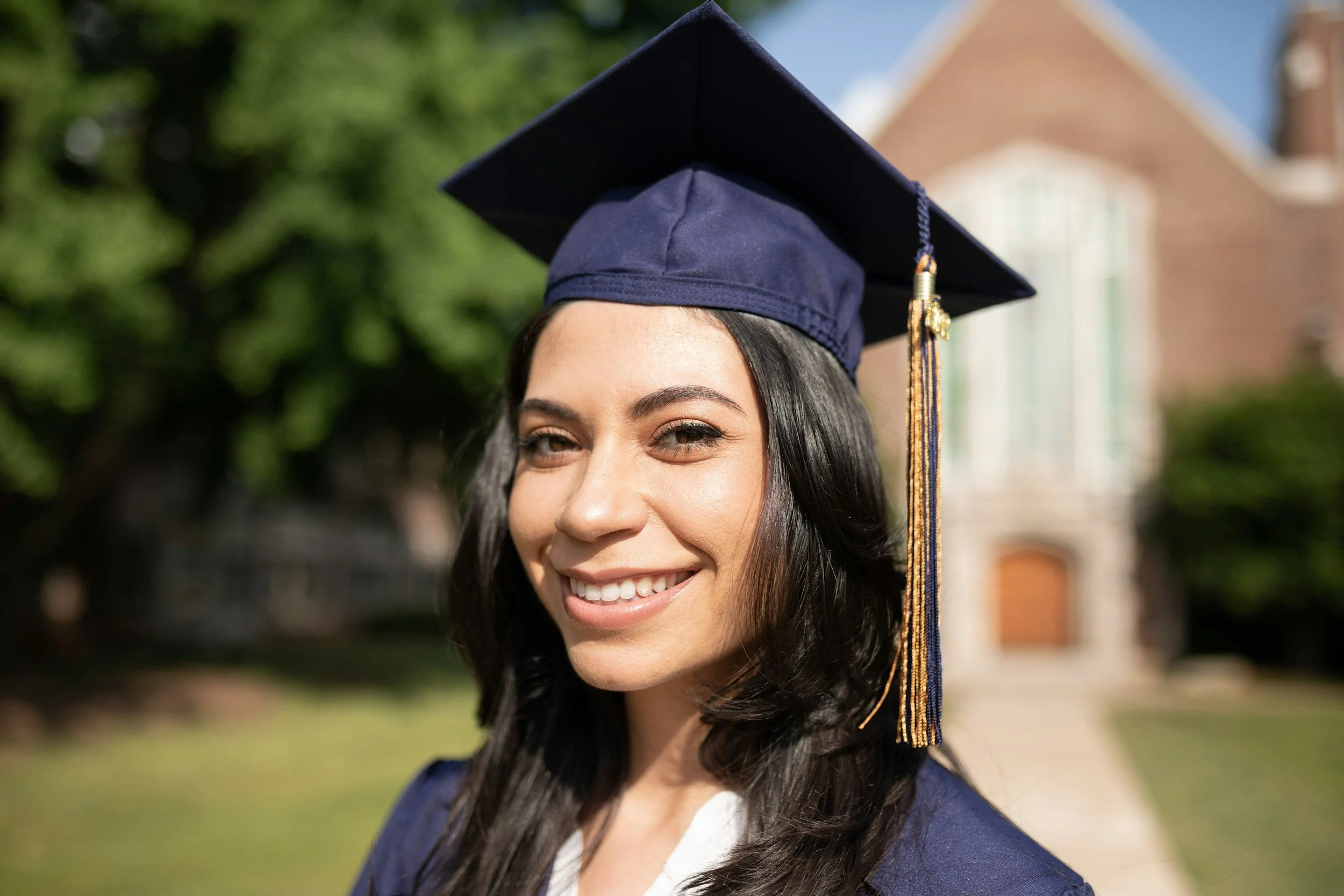 Young woman smiling in graduation cap and gown outdoors with greenery and a building in the background.