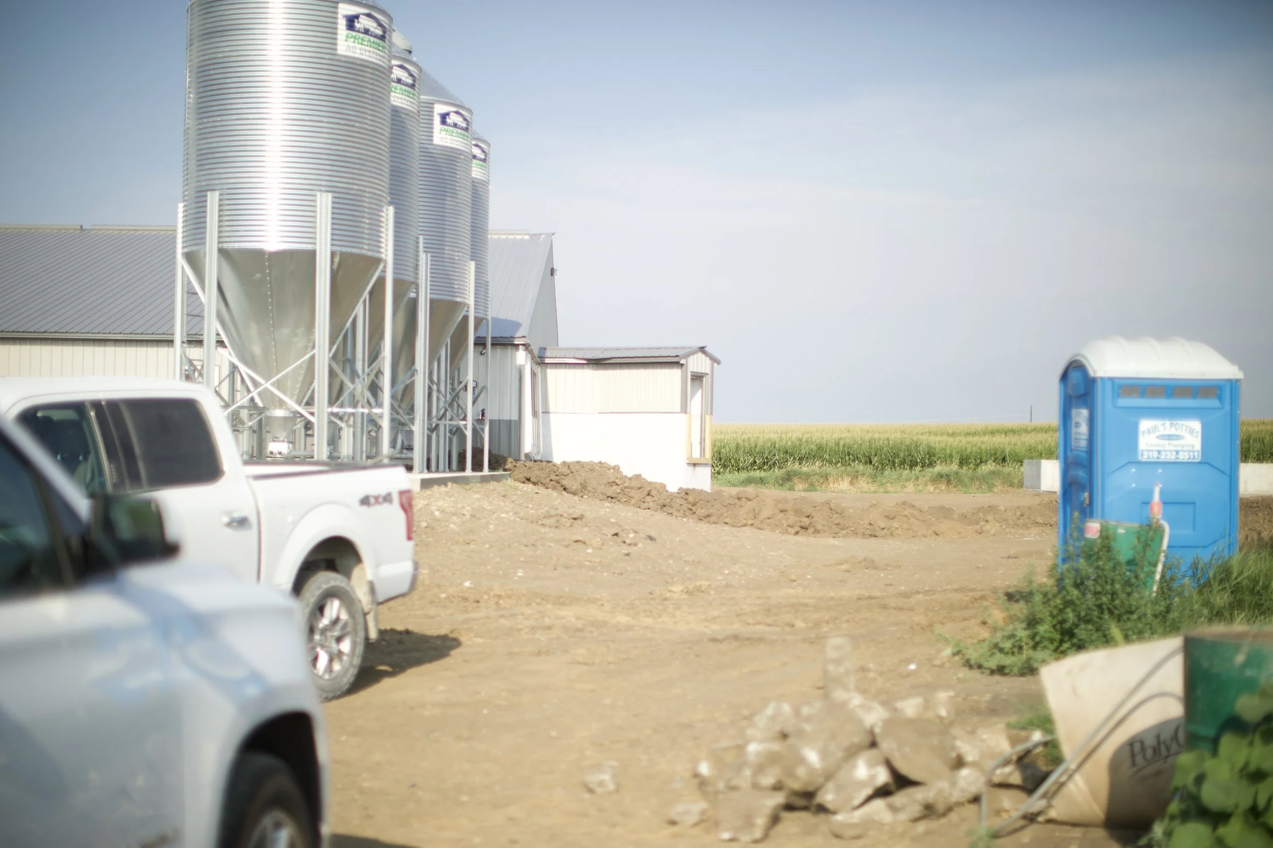 Farm or industrial site with silos, a building, and a portable toilet, with vehicles and construction materials in the foreground.
