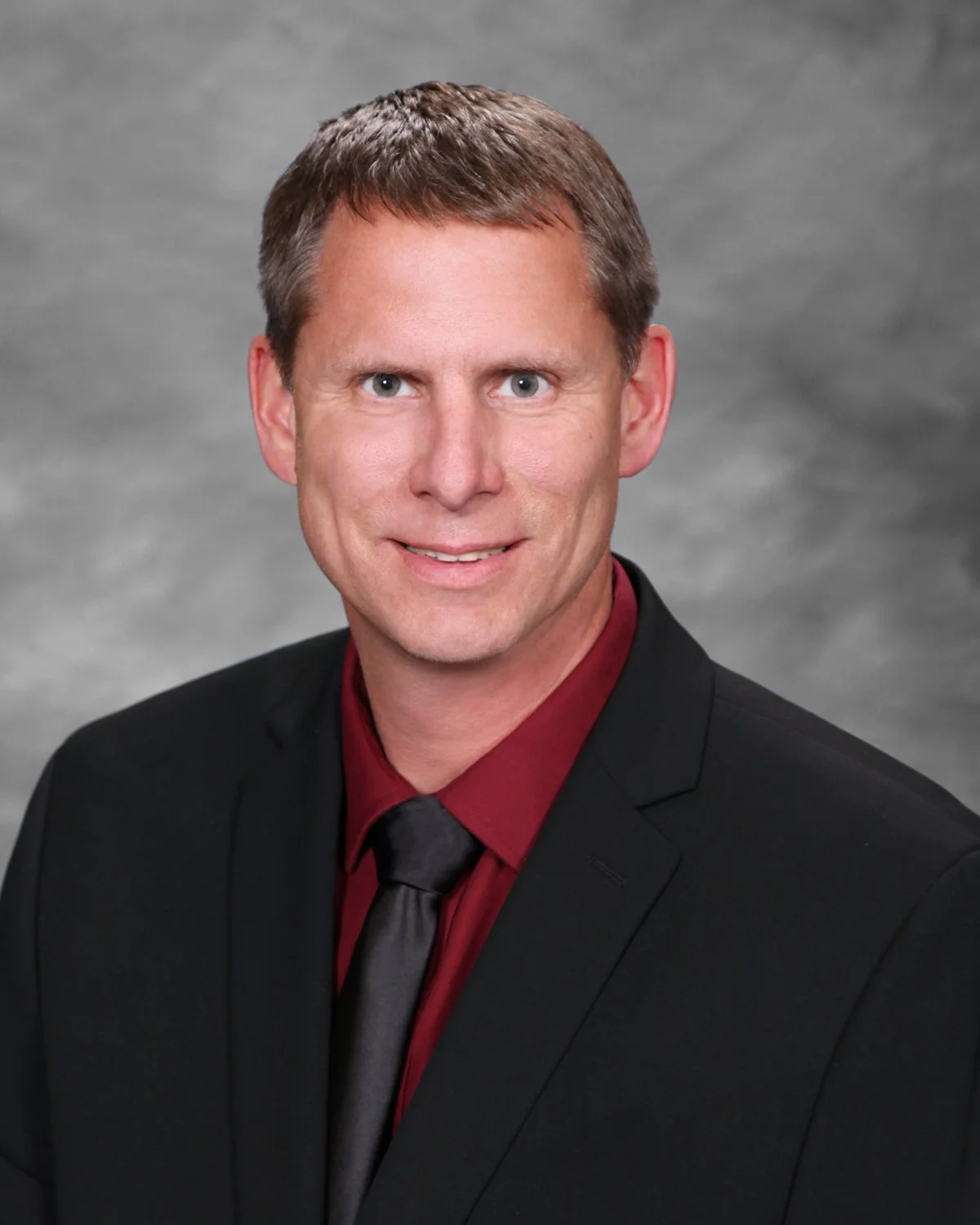 Headshot of a man with short brown hair wearing a black suit, red shirt, and black tie, against a gray background.