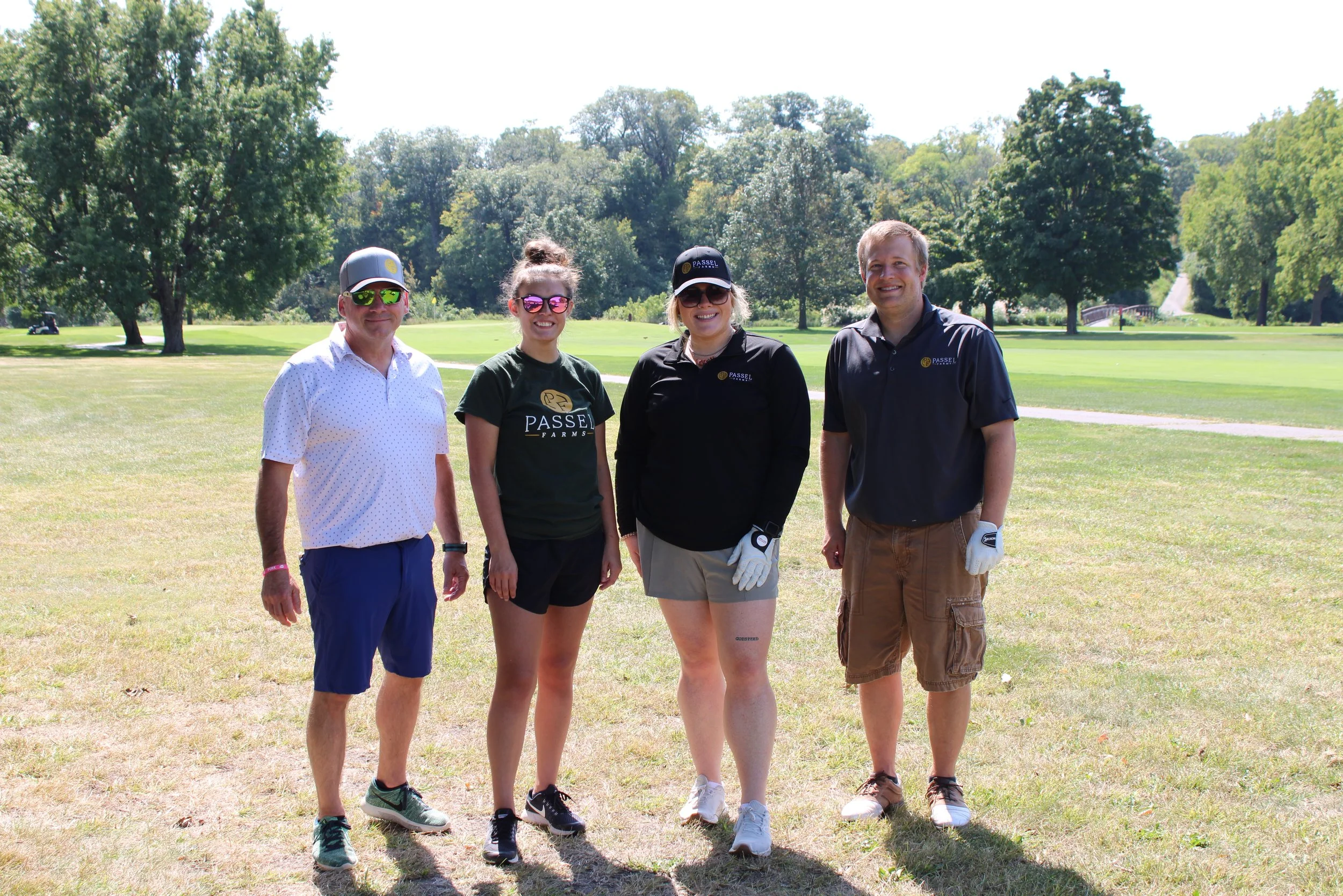 Four people standing on a golf course with trees in the background. They are smiling and dressed in casual golf attire, with some wearing sunglasses and caps.