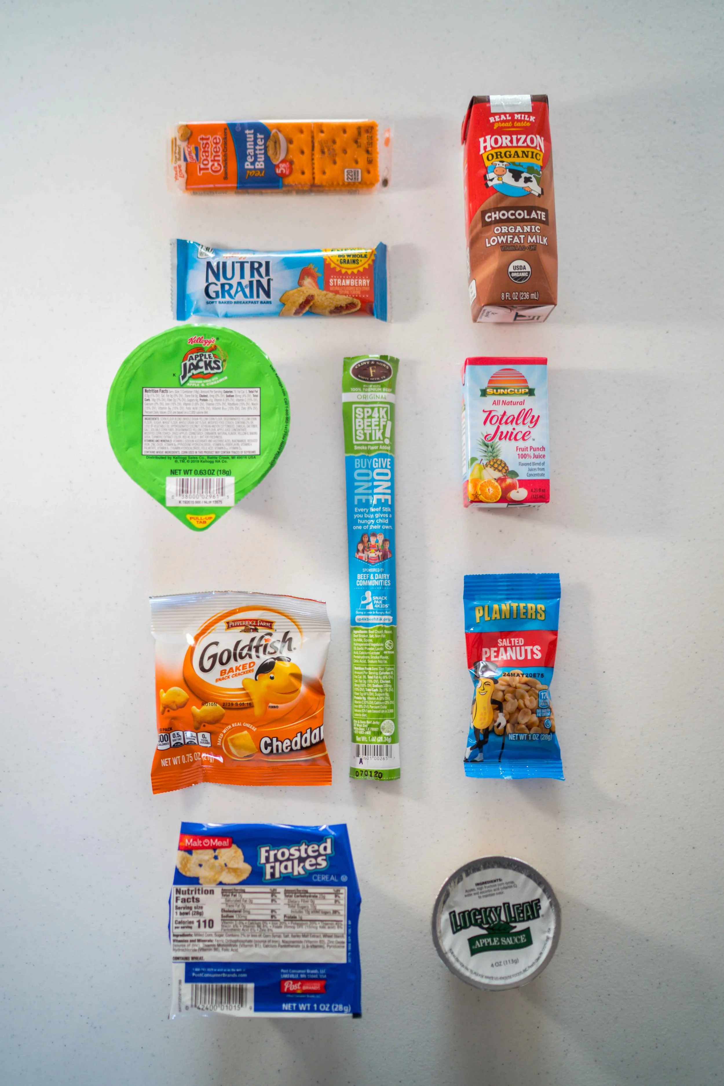 Array of grocery items on a white surface including a cheese danish, a green container of apple sauce, a box of frosted flakes cereal, a bag of salted peanuts, a roll of lunch meat, a chocolate low-fat milk carton, a strawberry nutri-grain bar, a string cheese package, a fruit punch juice box, and a snack cracker bag.