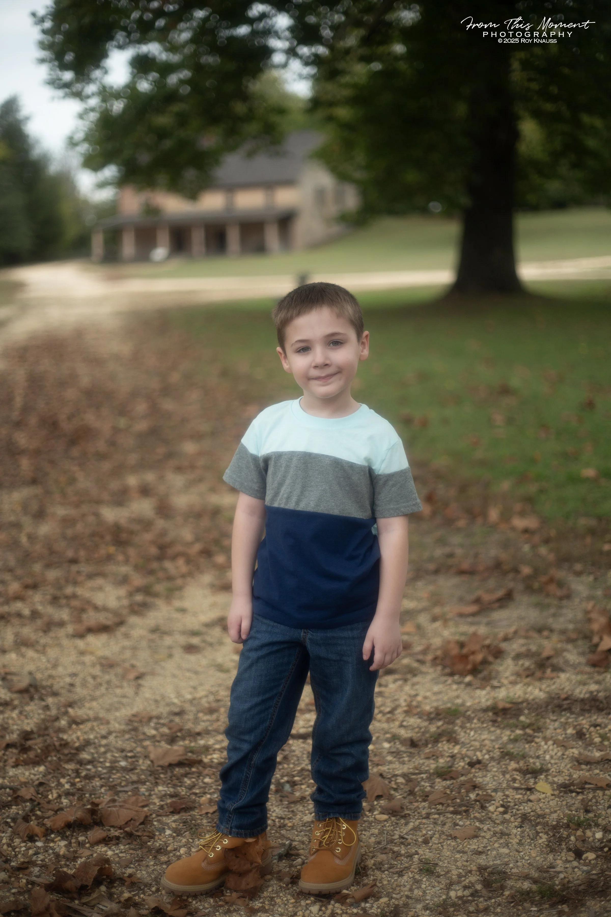 A young boy standing outdoors on a dirt and leaf-covered path, smiling at the camera with a house, large tree, and grassy yard in the background.