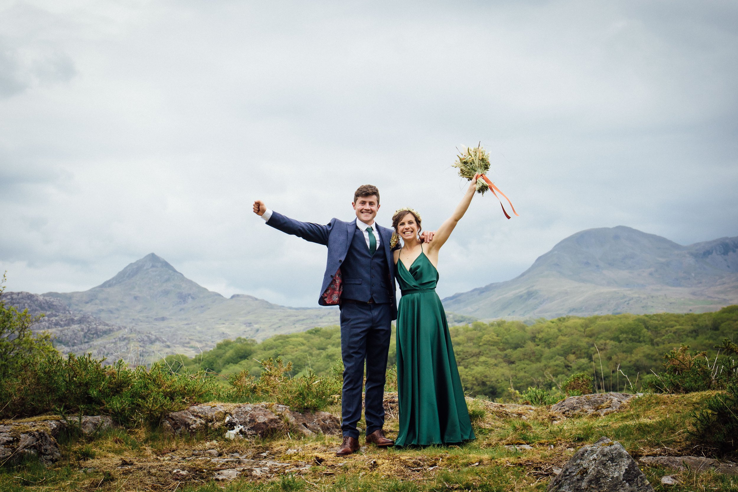 teleri and ned, owners of eryri barn events on their wedding day with arms up in the air, teleri wears dark green dress and ned wears a blue suit