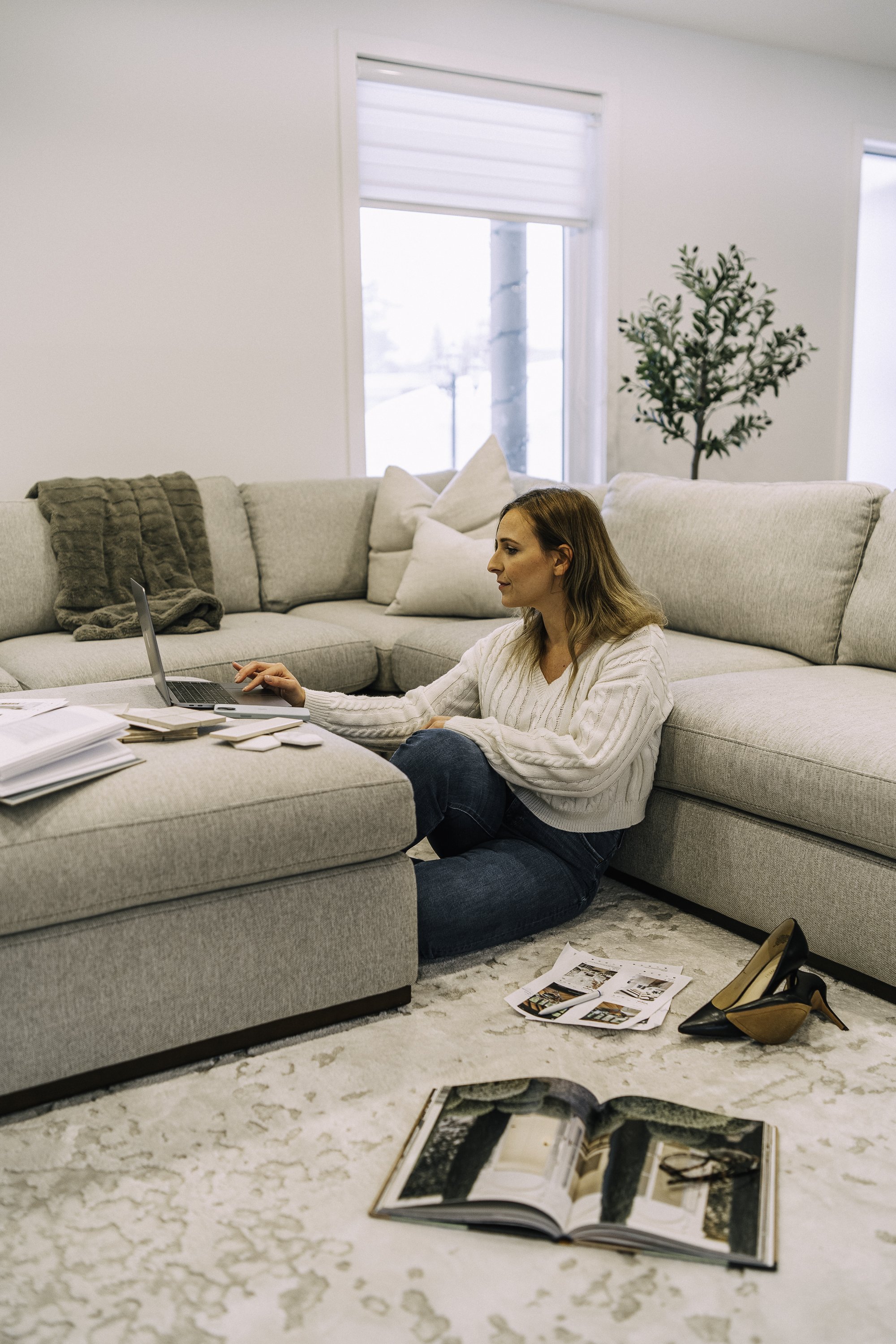 A woman sitting on the floor beside a large beige sectional sofa, working on a laptop with various papers and magazines scattered around her in a bright living room with large windows and a tall plant.
