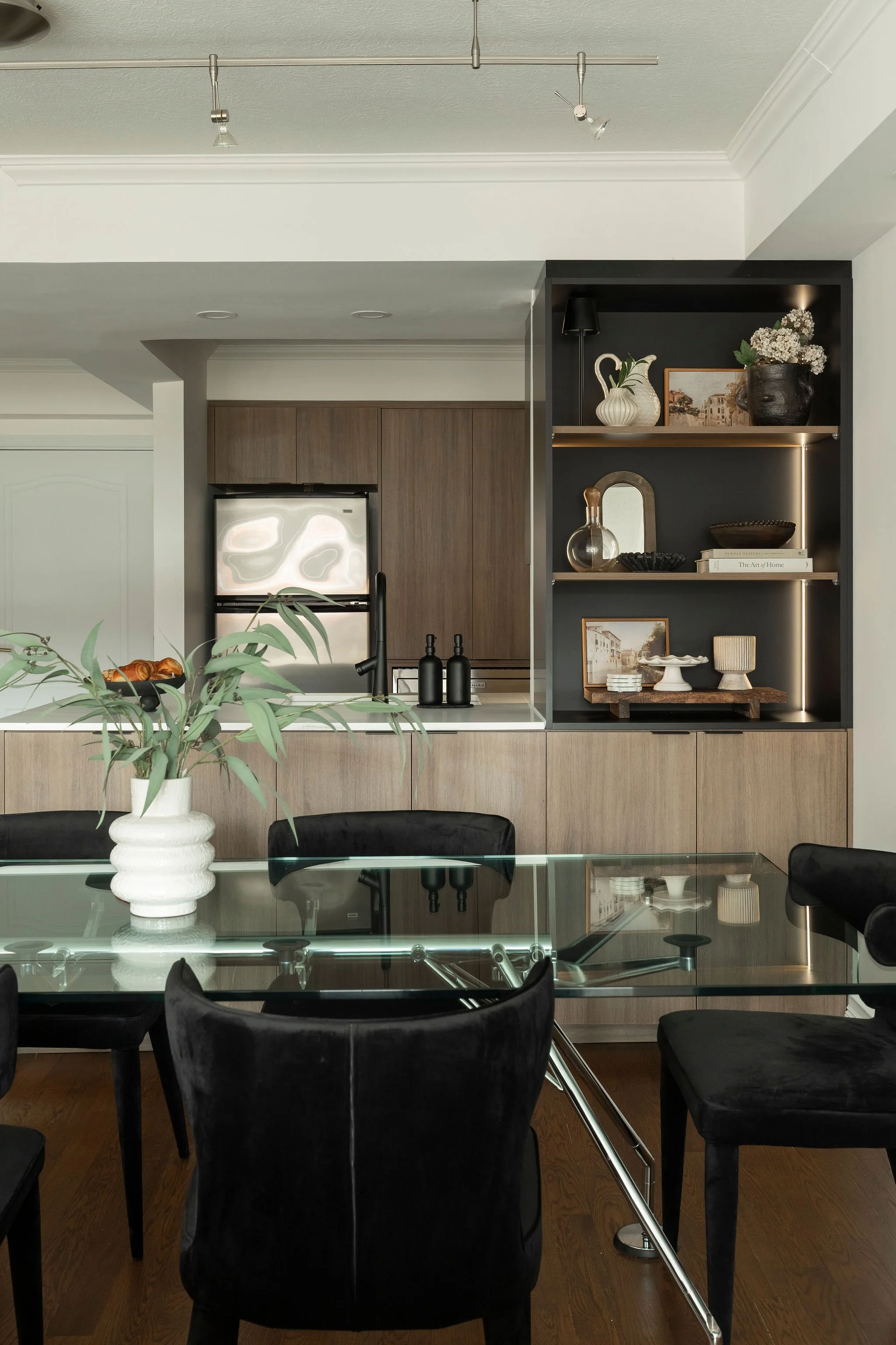 Modern dining room with a glass-top table, black chairs, a potted plant in a white vase, and a kitchen in the background featuring wooden cabinets, a stainless steel refrigerator, and decorative shelving with vases, framed art, and books.