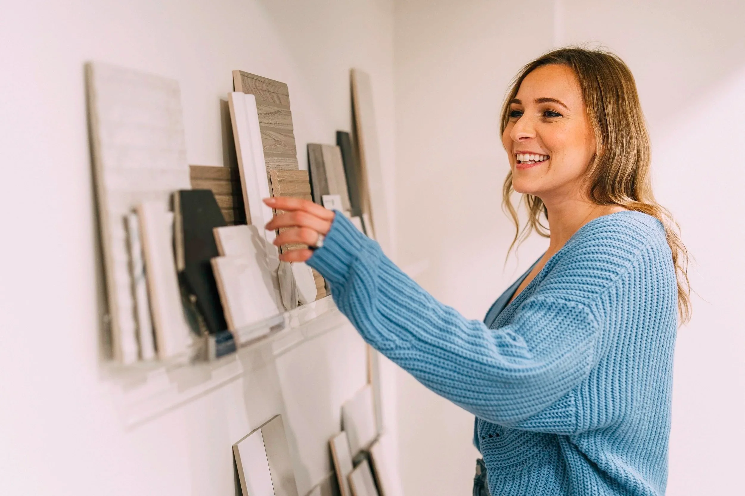 A woman with shoulder-length blond hair wearing a blue knitted sweater smiling while pointing at wall-mounted display samples of wood and tile options.