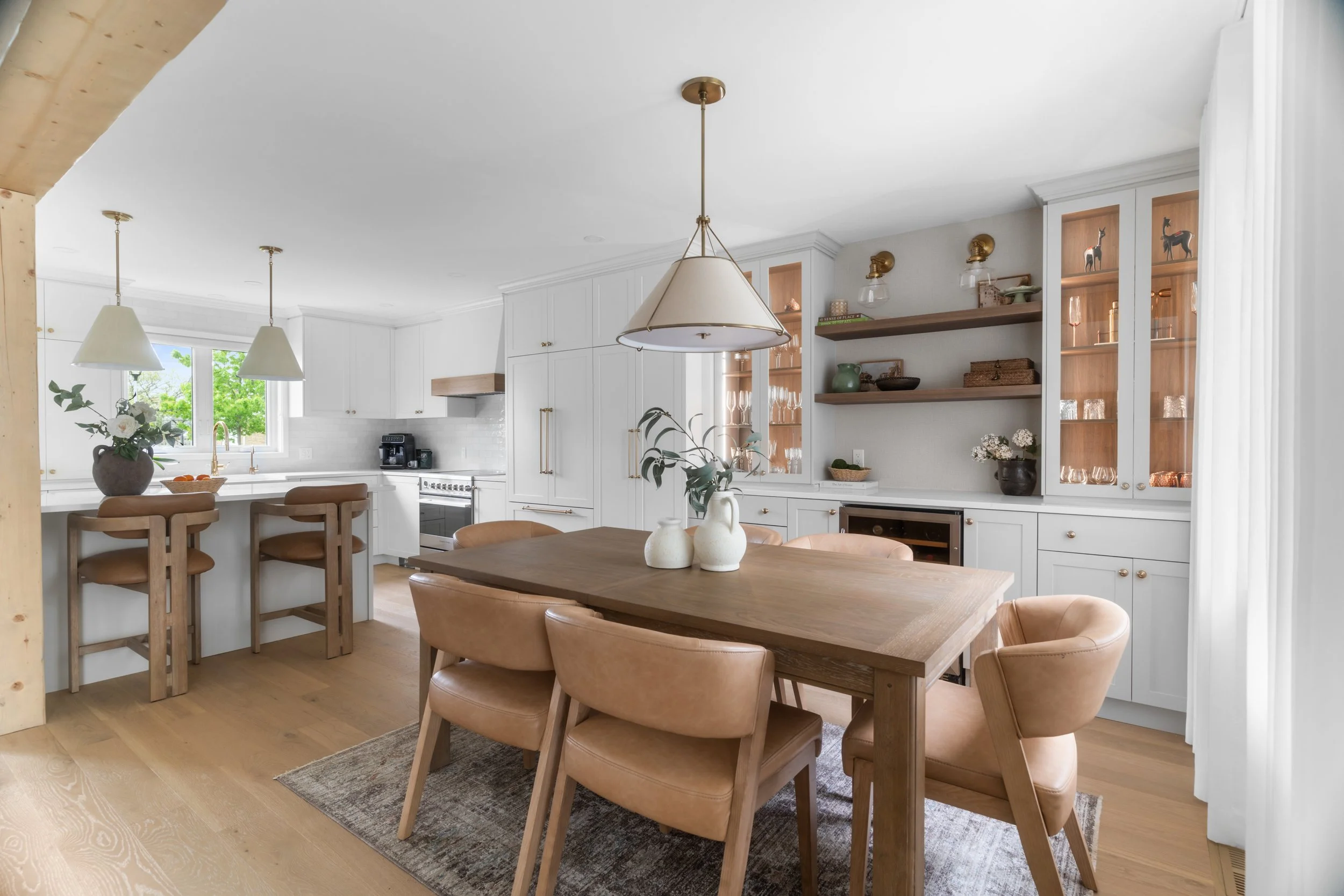 Open-concept kitchen and dining area with white cabinets, wooden accents, and beige leather chairs around a wooden dining table. Pendant lights hang above the kitchen island, and decorative shelves display glassware and ornaments.