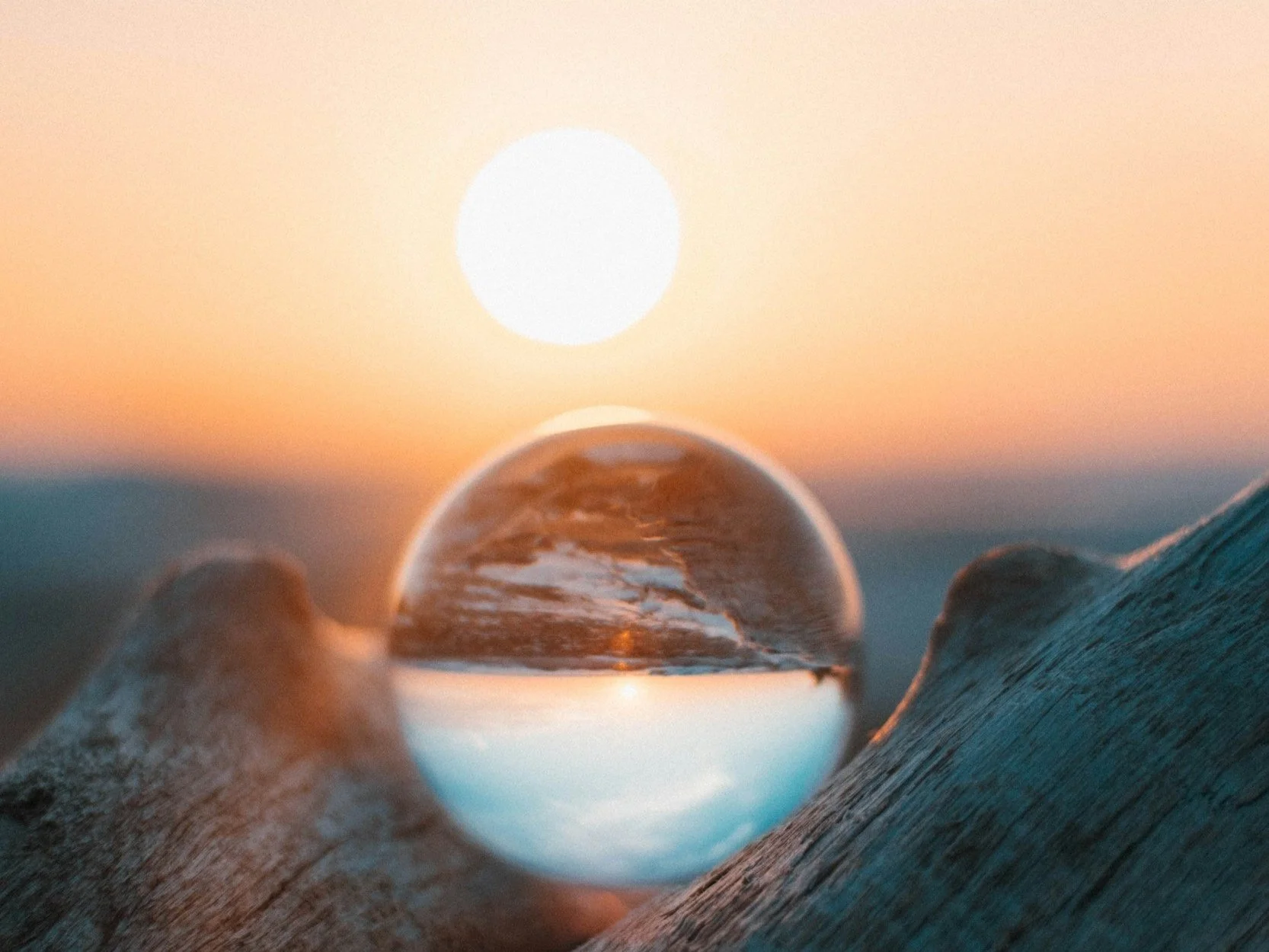 Nahaufnahme eines transparenten Glasballs, der eine reflektierte Meereslandschaft mit Wellen und Himmel zeigt, auf einem Holzstück bei Sonnenuntergang.