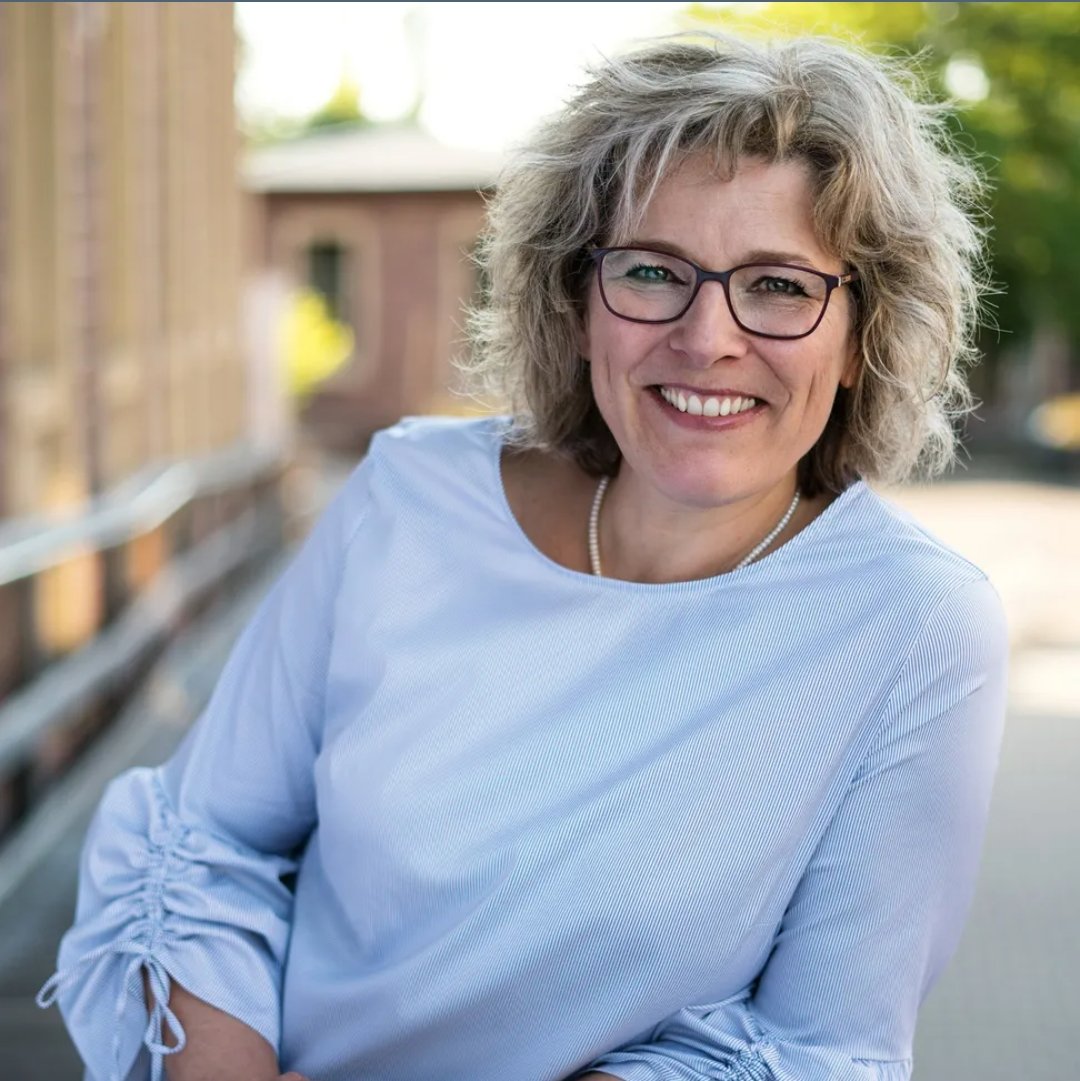 Eine lachende Frau mit Brille und lockigem, grauem Haar, trägt ein weißes Oberteil, sitzt draußen auf einer Bank mit Natur im Hintergrund.