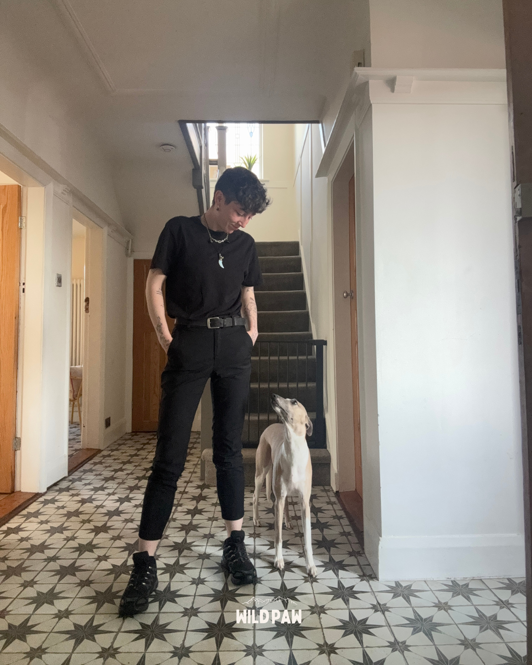 A person with short black hair, tattoos, and dressed in black pants and t-shirt, standing on a patterned tiled floor inside a home, looking at a tall, tan and white dog. The person is wearing black shoes and has hands in their pockets, while the dog is looking up at them.