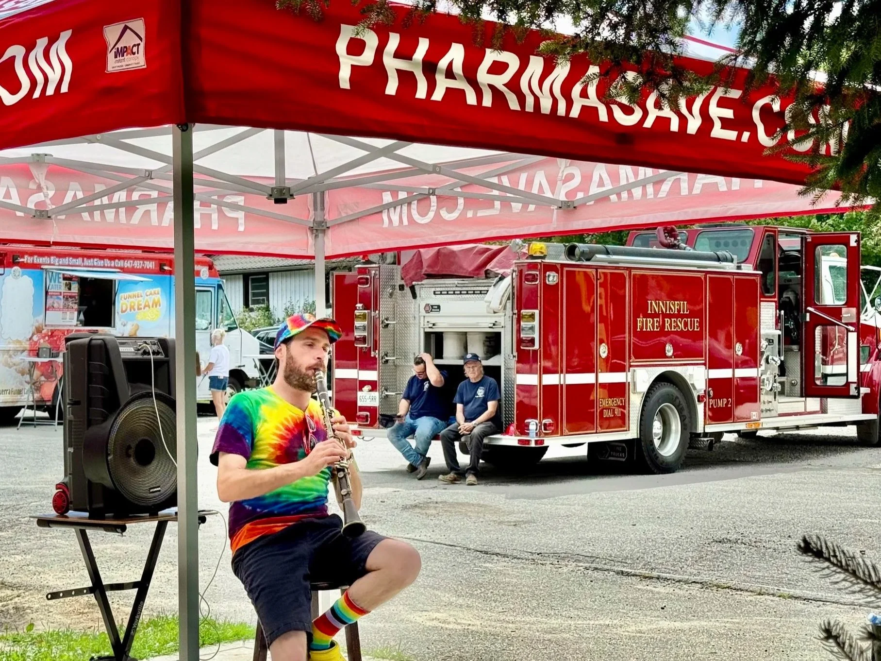 A man in a rainbow tie-dye shirt and rainbow socks playing a clarinet at an outdoor event. Behind him, there is a red fire truck from Innissfil Fire Rescue and a small group of people sitting on chairs. There is a red canopy and a food truck in the background.