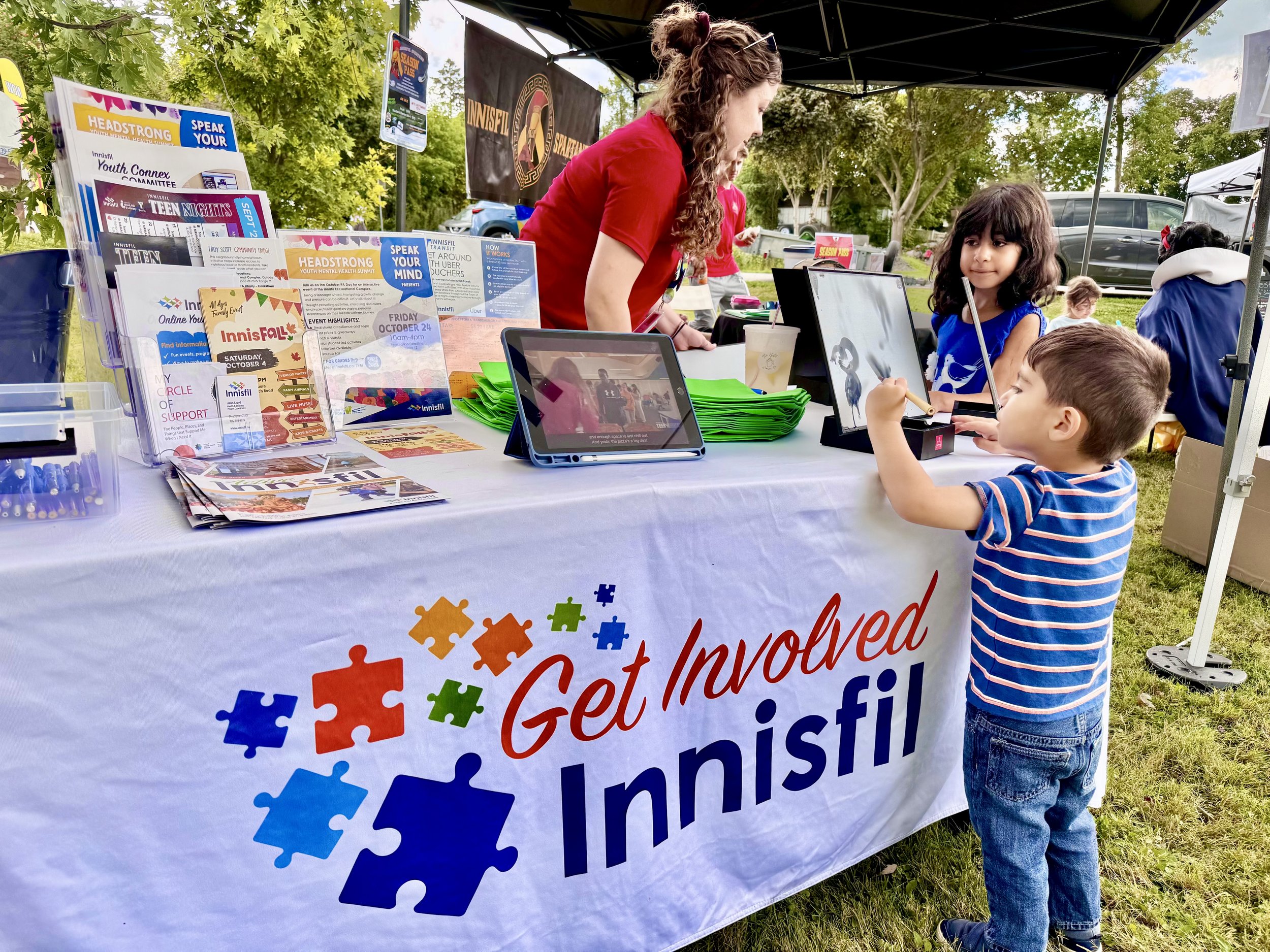 Child in striped shirt and jeans at a booth with pamphlets and tablets, outdoor event surrounded by trees.