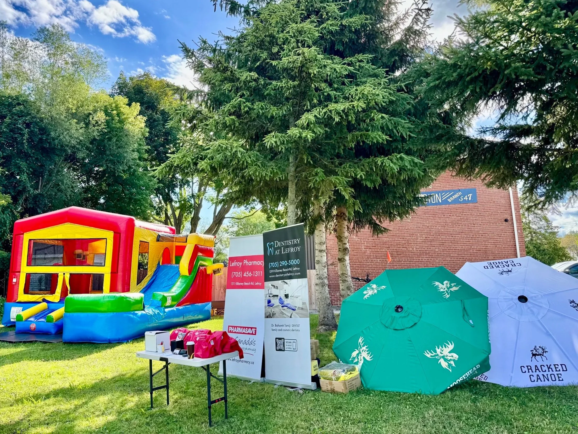An outdoor community event with a colorful inflatable bounce house, tents, and signs for local businesses like dentistry and pharmacy. Green trees and a blue sky are visible.