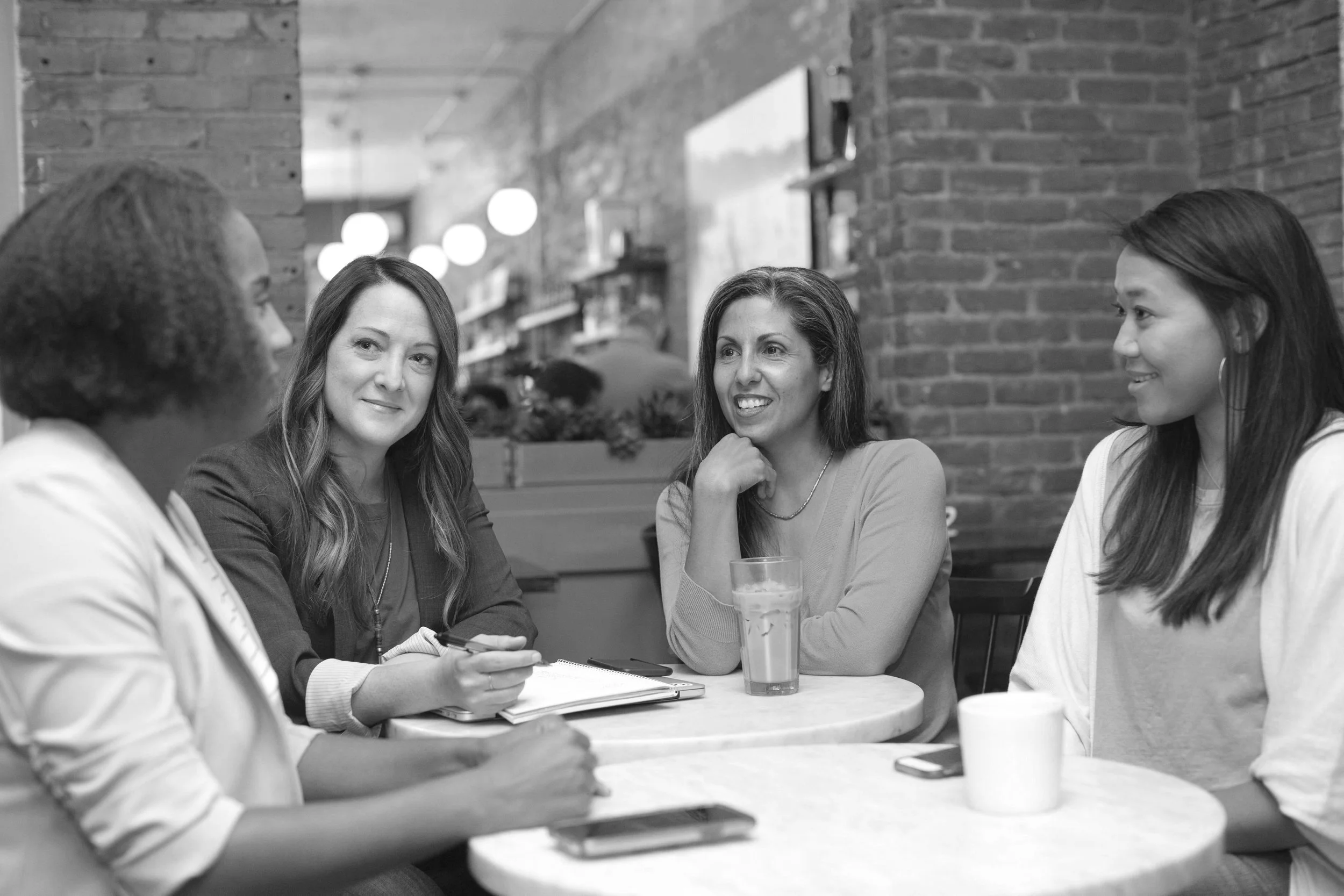 Four women sitting around a table having a conversation in a cafe with brick walls and hanging lights.