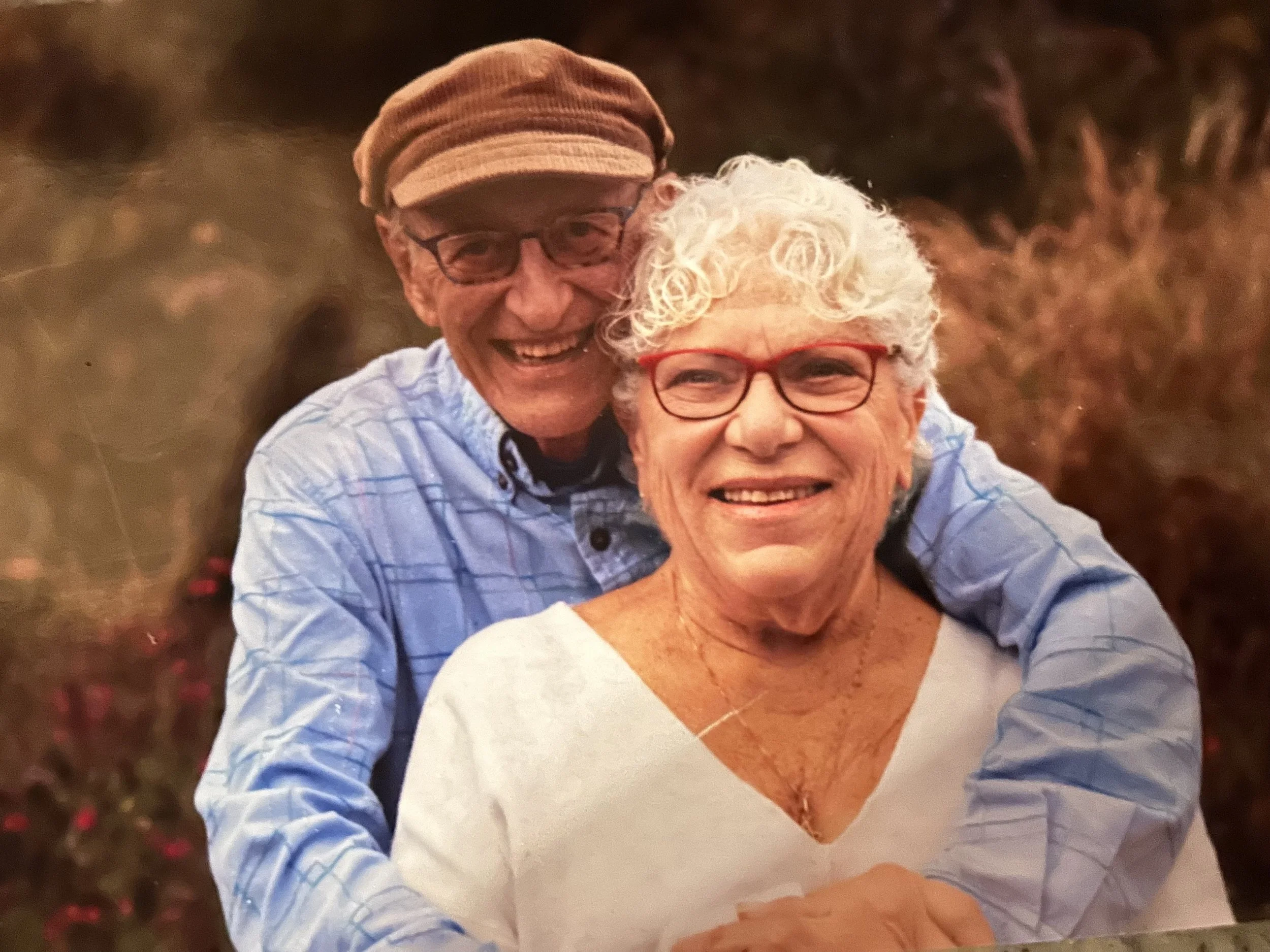 An elderly man and woman smiling and hugging outdoors, with a blurred natural background.