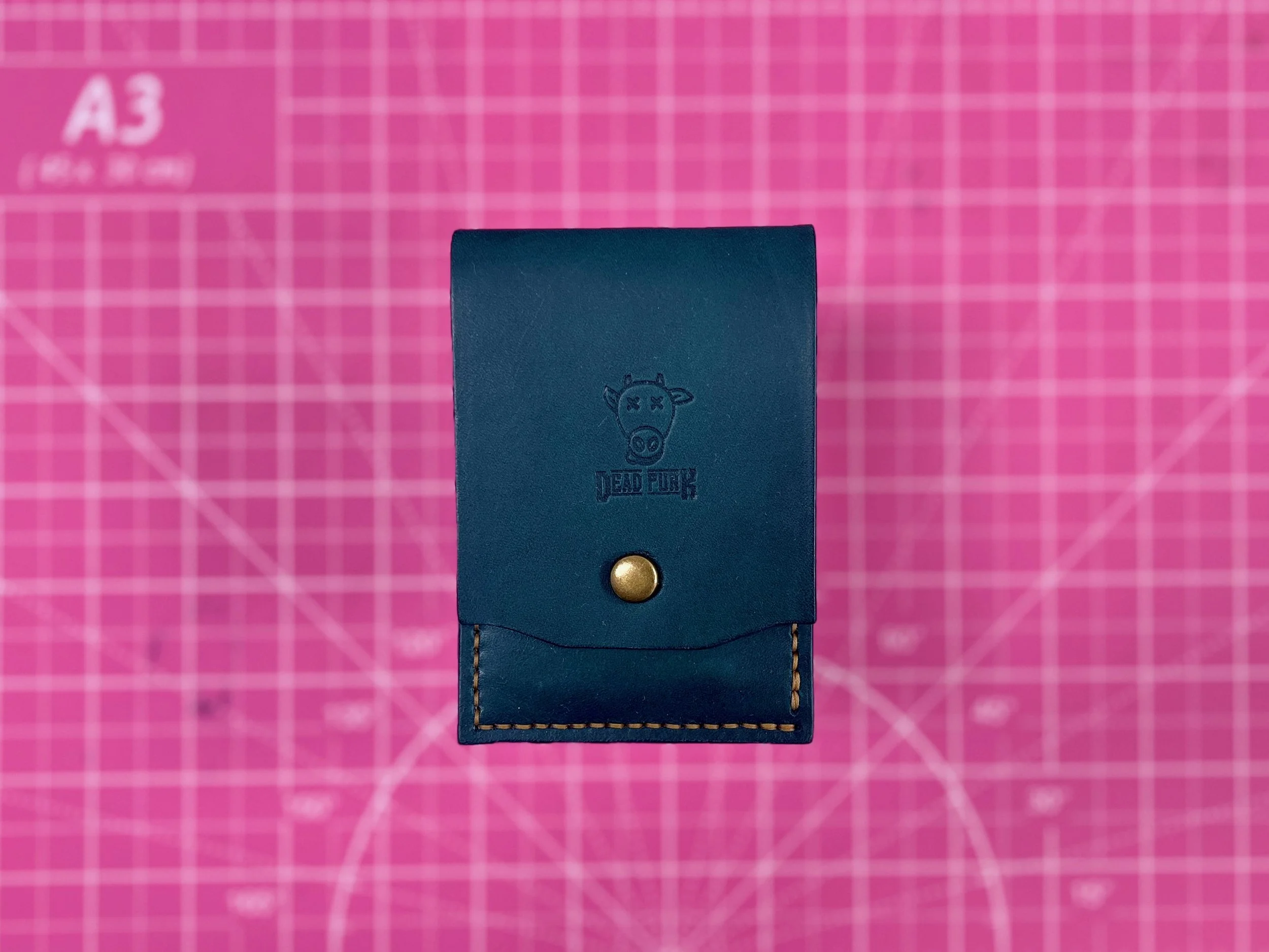 A handcrafted deep teal leather pocket pouch with a brass snap closure and the Dead Punk Leather cow logo embossed on the front flap, resting on a pink cutting mat.