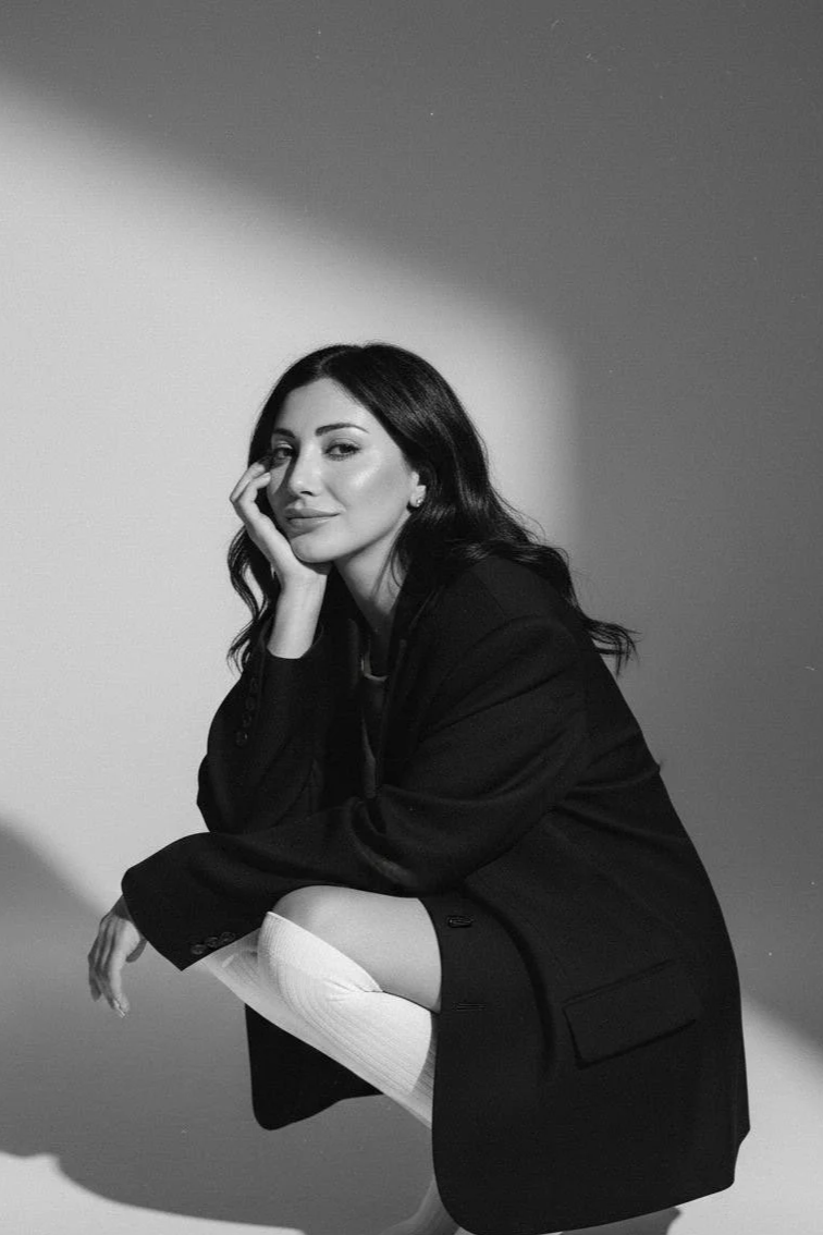 Black and white photo of a woman with dark wavy hair, sitting with one knee up, wearing an oversized blazer and thigh-high stockings, looking at the camera with a relaxed expression.